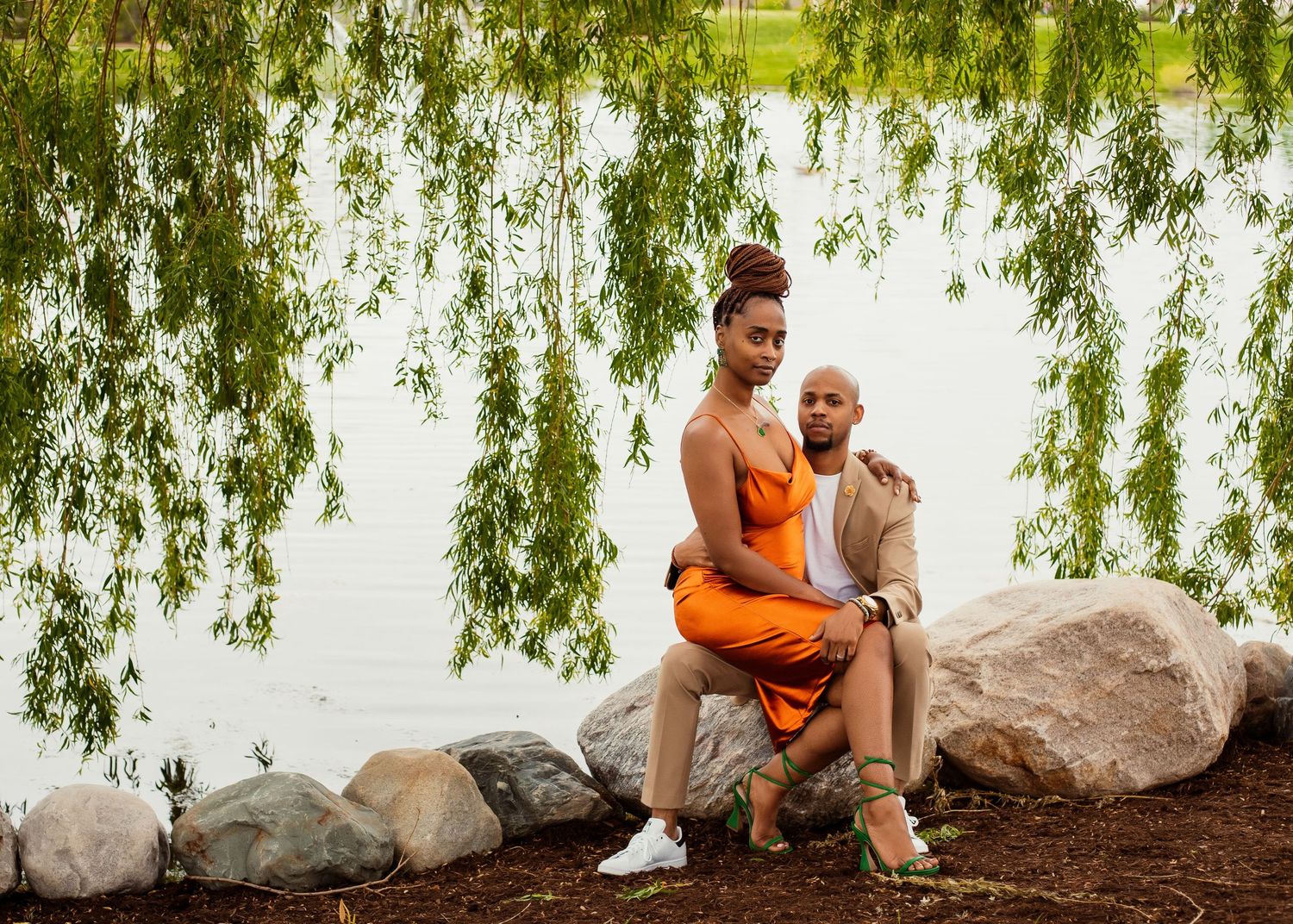 Couple sitting on rocks by the lake, wearing orange dress and beige suit under weeping willow branches.