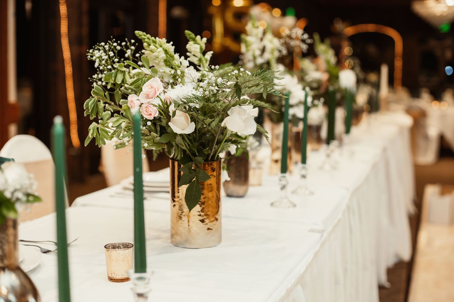 Elegant floral centerpieces with white flowers and greenery line a long reception table.