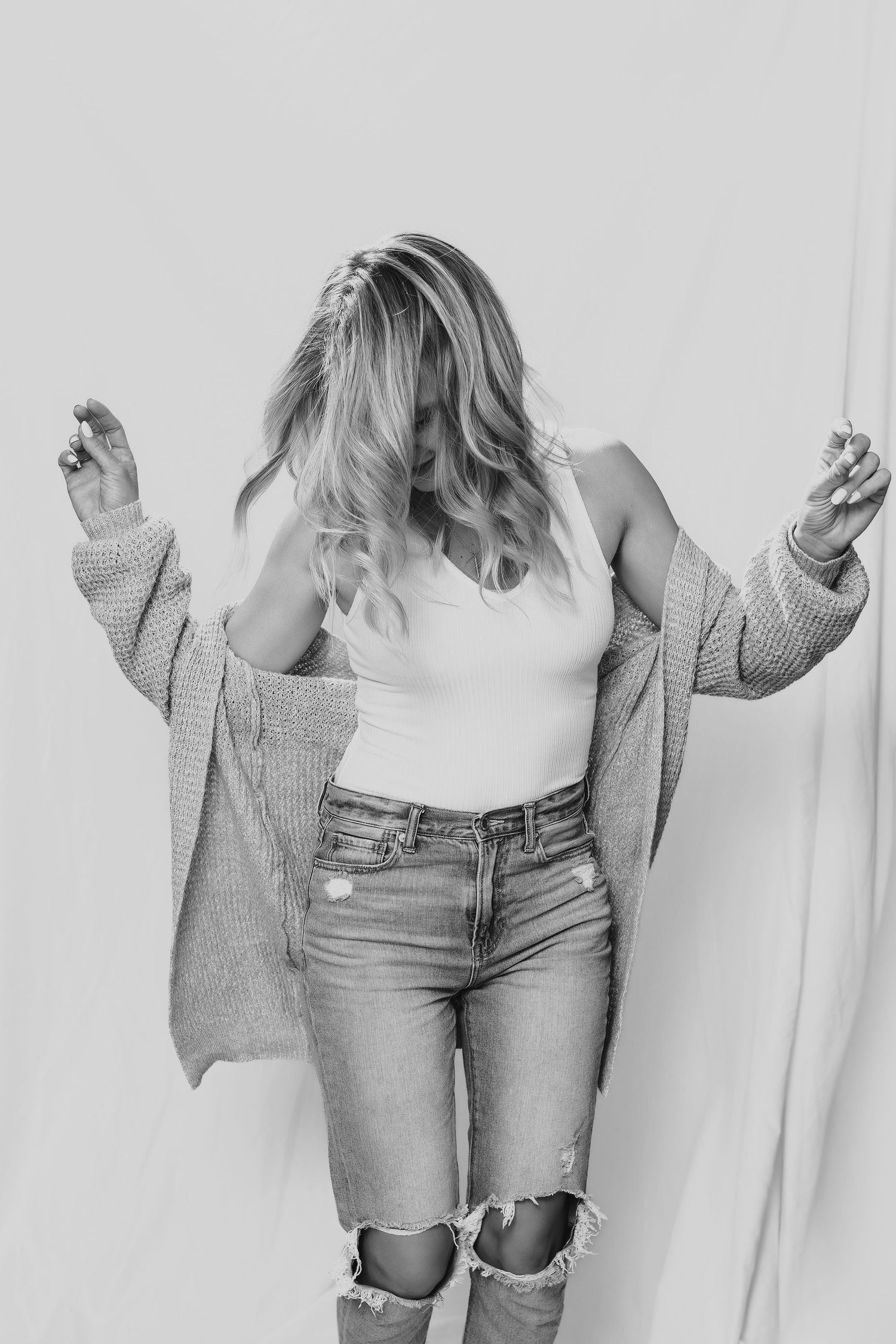 Black and white portrait of someone in distressed jeans, white tank top and oversized knit cardigan against light backdrop.