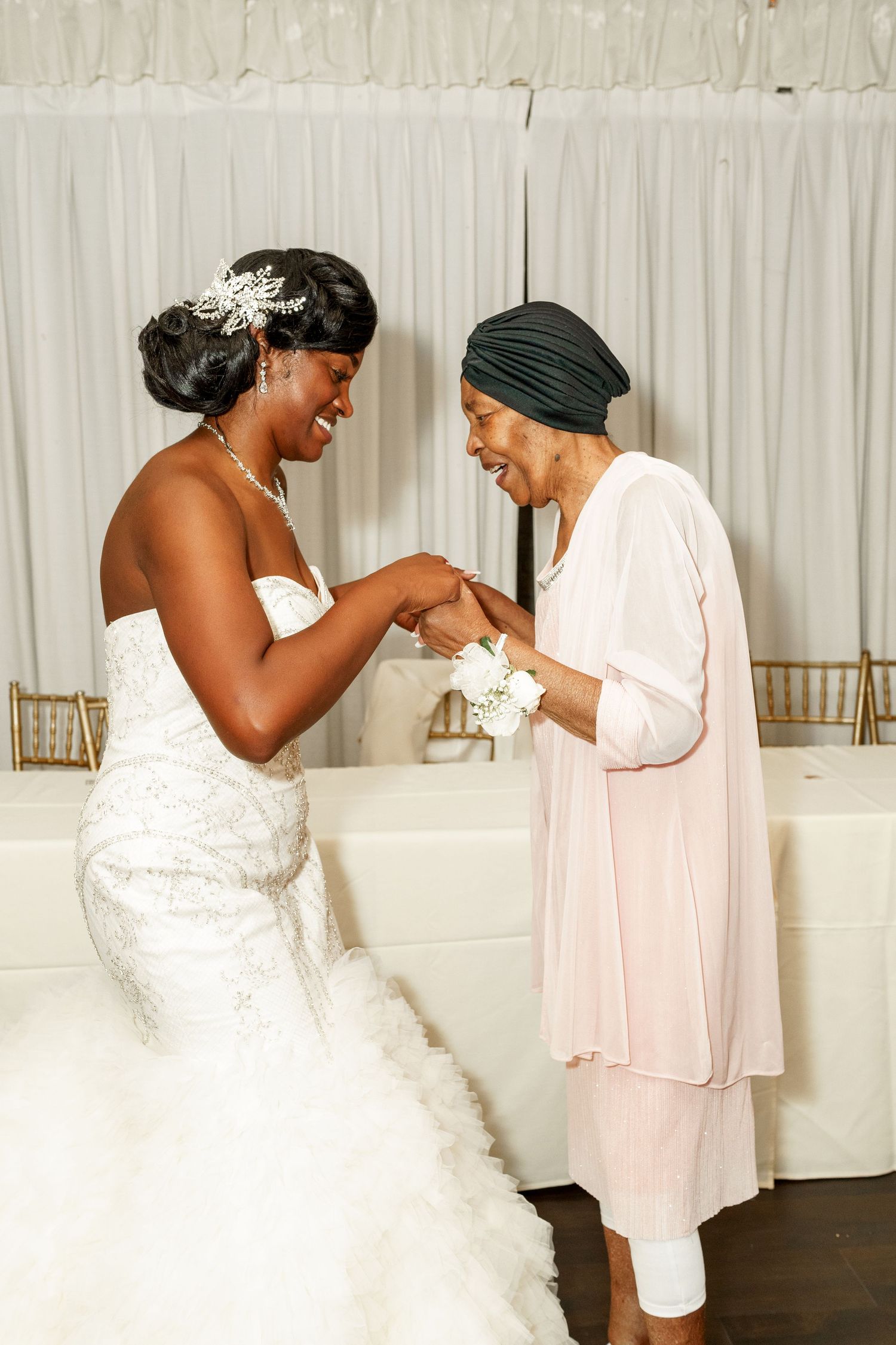 A joyful moment shared between the bride in a white mermaid gown and an elder wearing a light pink outfit at a wedding.