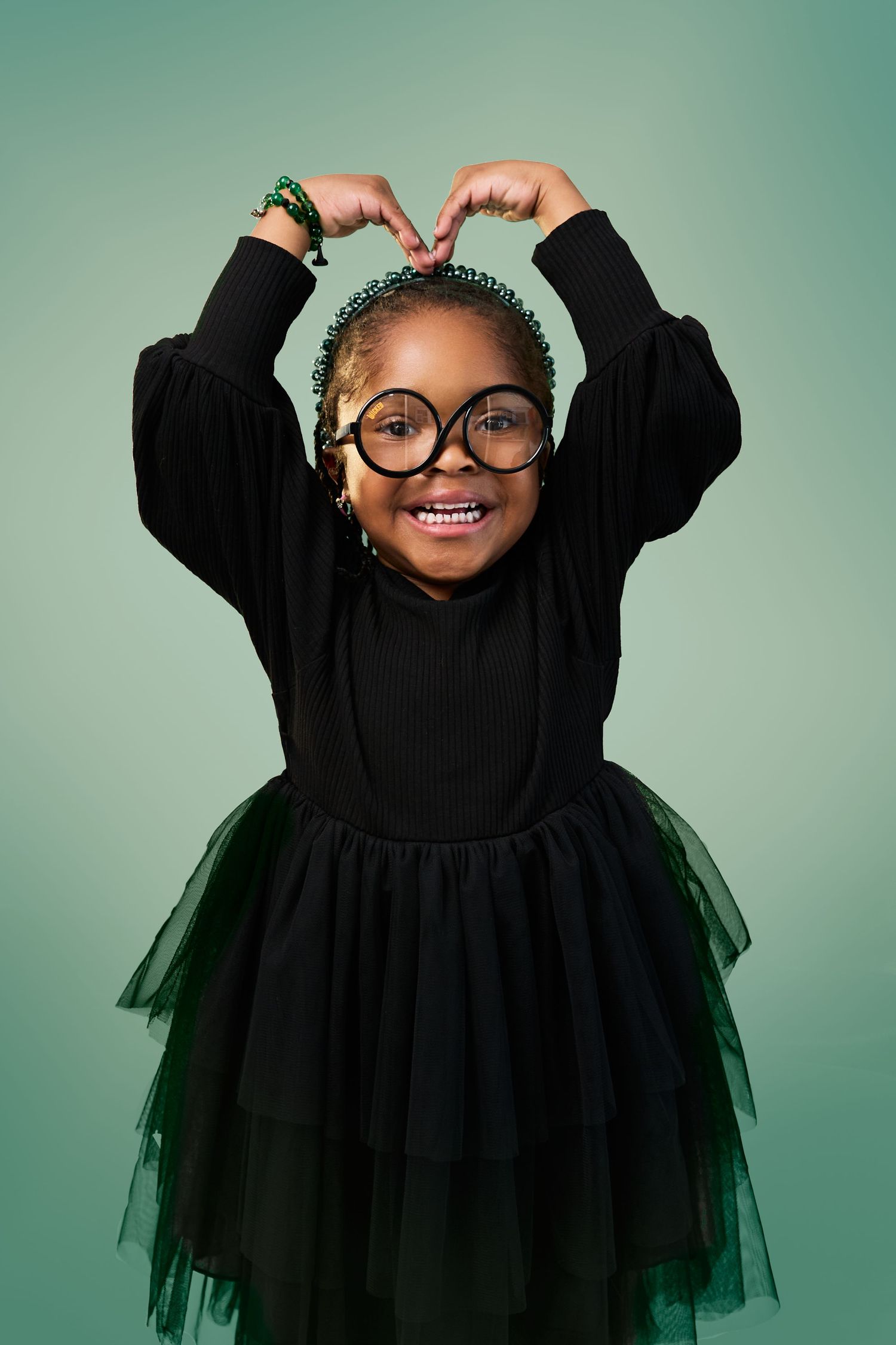 A child in glasses and a black tulle dress poses playfully against a teal background with arms raised and joyful expression.