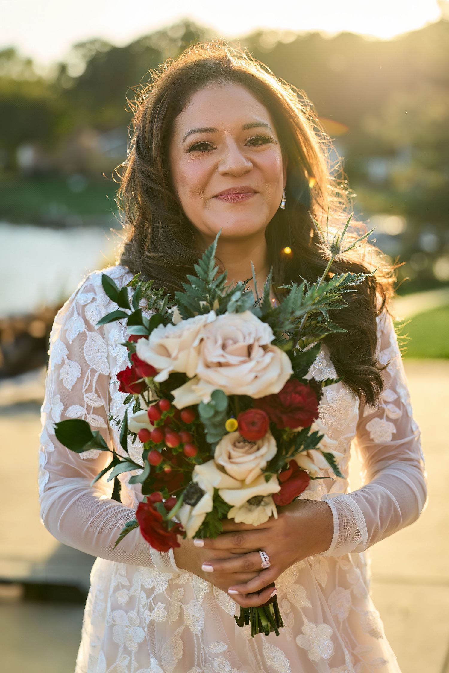 A bridal bouquet with white and red flowers is held in golden sunlight against a blurred outdoor background.