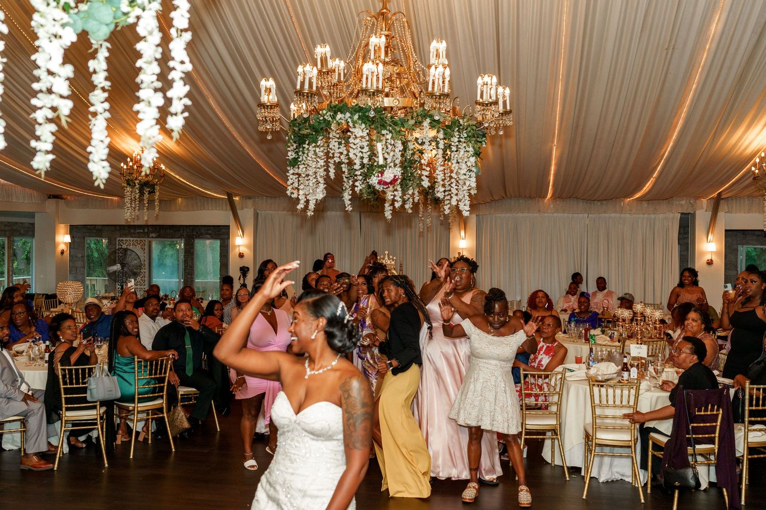 Wedding celebration with guests dancing under elegant chandeliers and draped fabric ceiling.