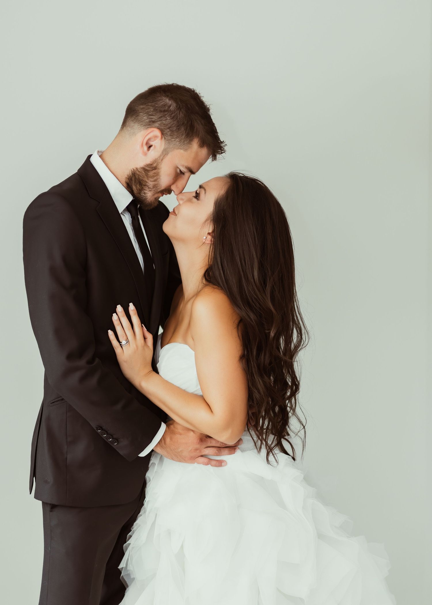 An intimate wedding portrait of a couple in formal attire sharing a tender moment against a neutral wall.