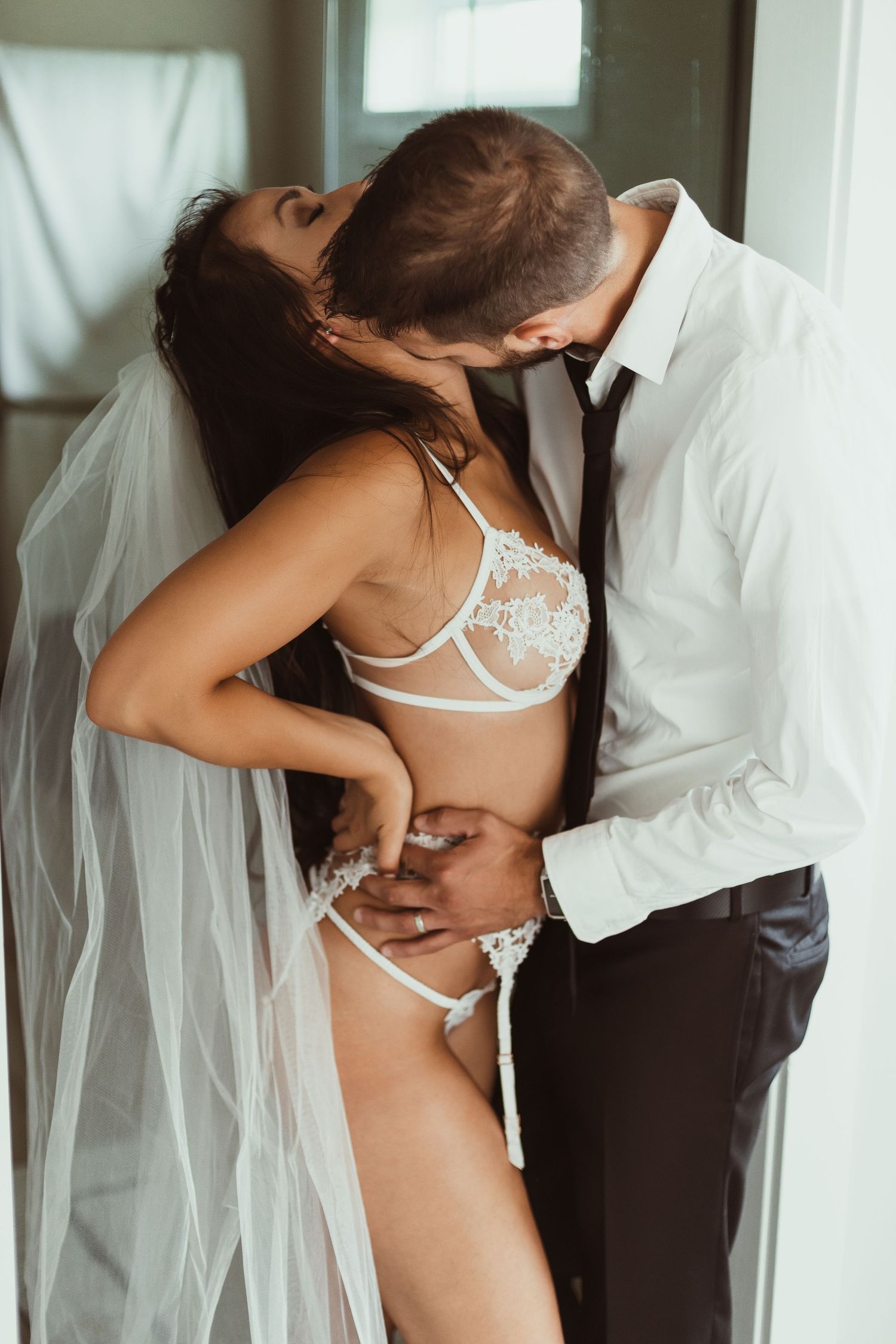 Romantic bridal boudoir photo of couple embracing in white lingerie and formal wear.