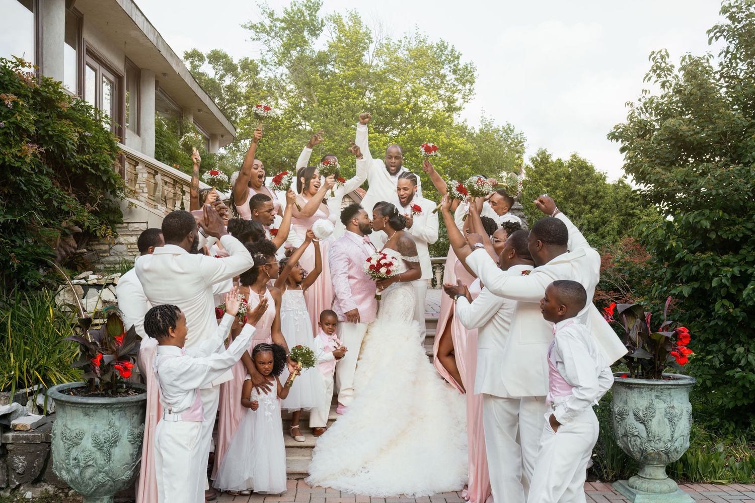 Large wedding party in white and pink attire celebrates enthusiastically on outdoor stone steps.
