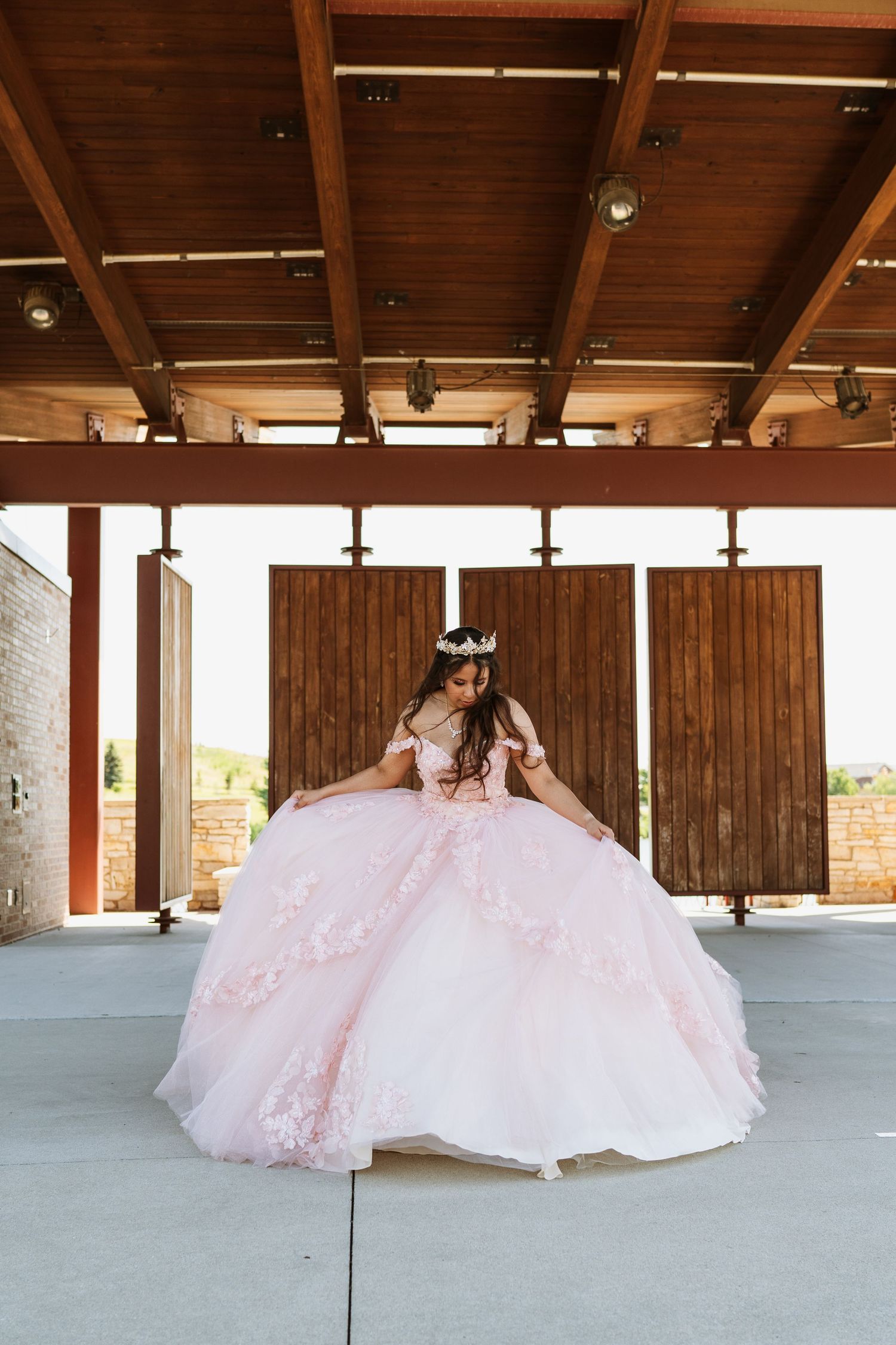 Pink princess ball gown dress with full tulle skirt photographed under wooden beamed pavilion on concrete floor.