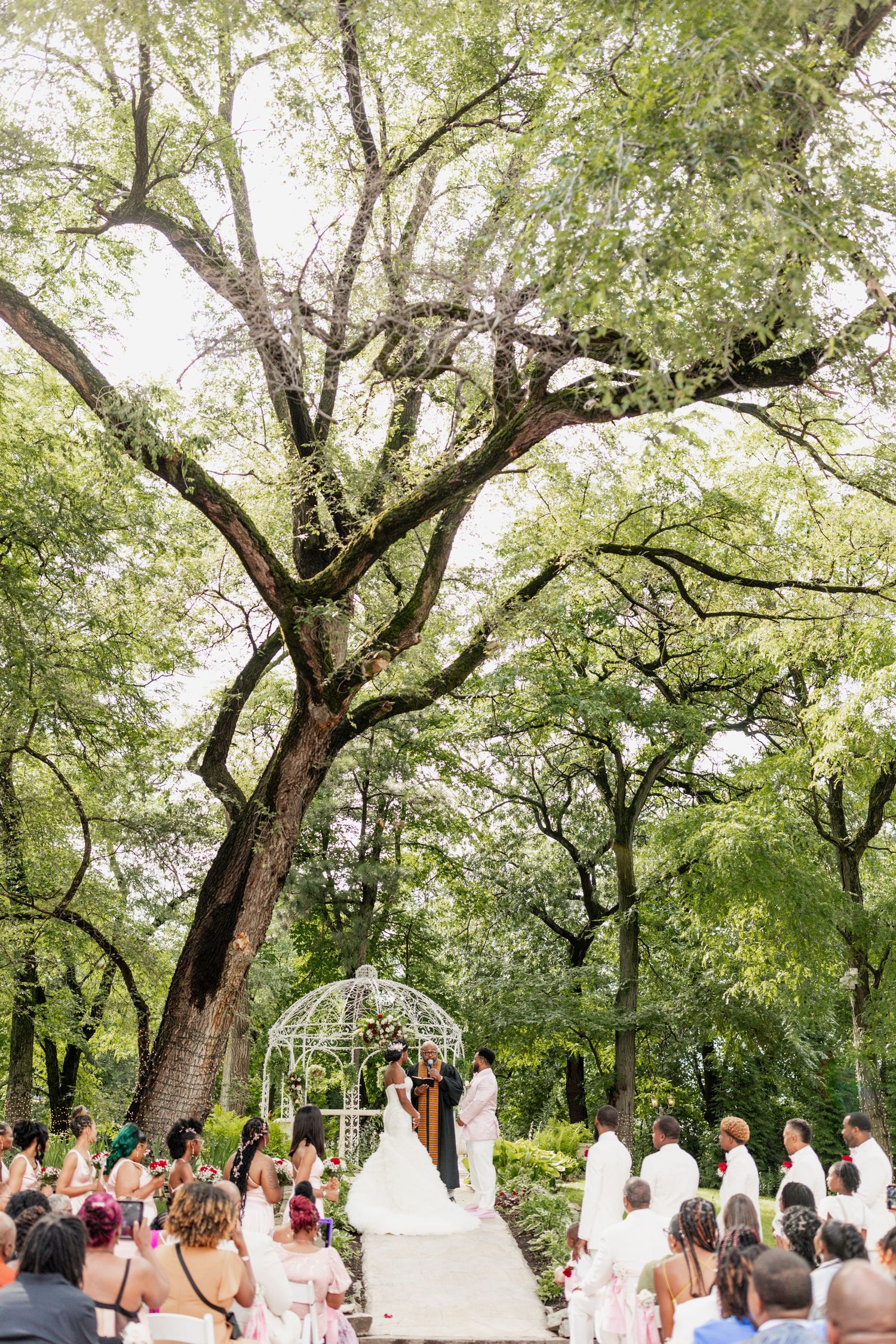 A wedding ceremony takes place under large trees with a white gazebo and guests seated on both sides.