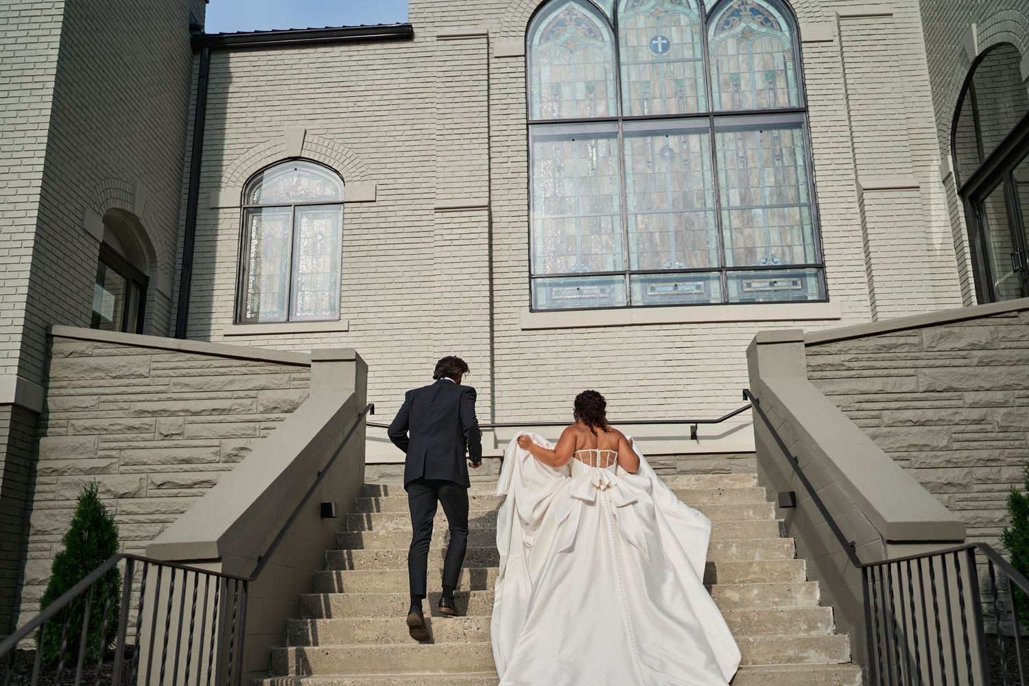 A couple walking up the grand stone steps of a church with ornate arched windows.