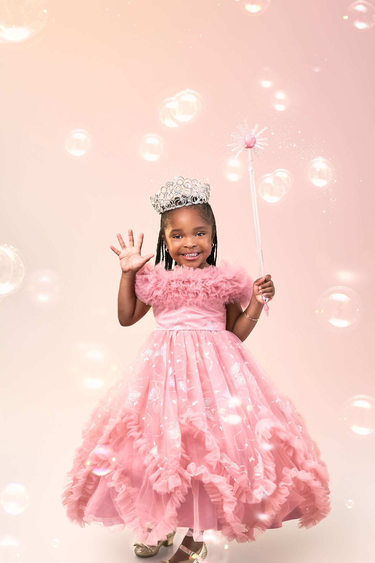 Child in pink princess dress waves and holds wand with bubbles floating in magical pink backdrop.