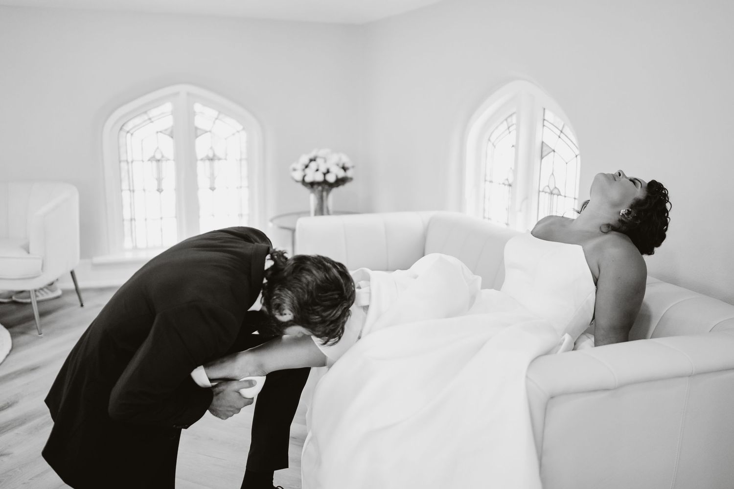 A black and white photograph of two figures in formal attire sharing an emotional moment in a white room with arched windows.