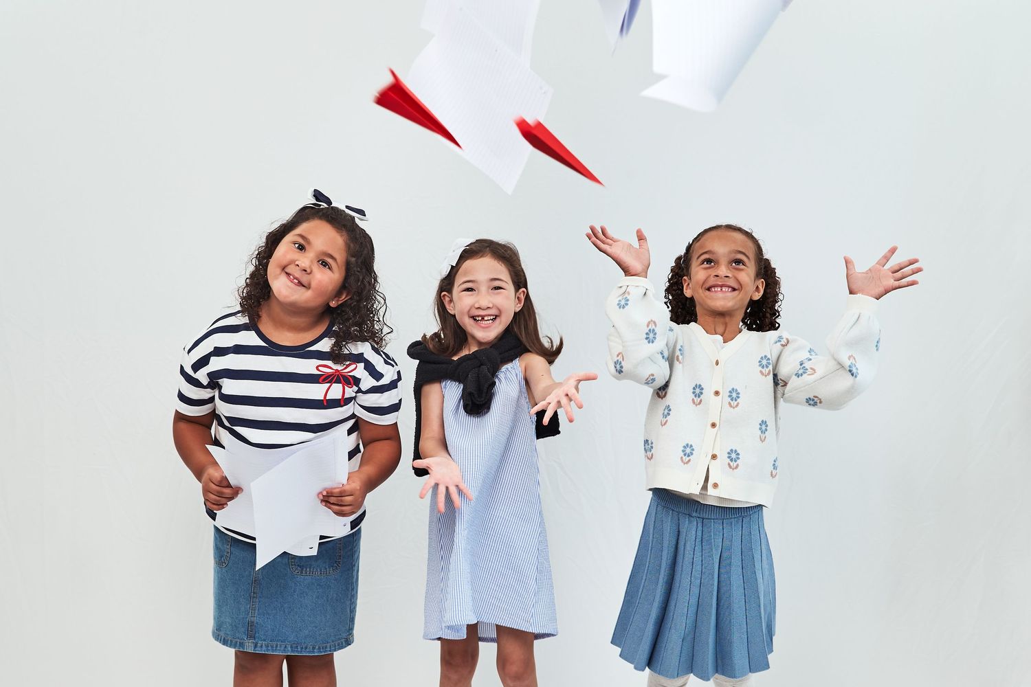 Three excited children in casual outfits standing against white wall throwing paper planes in the air.