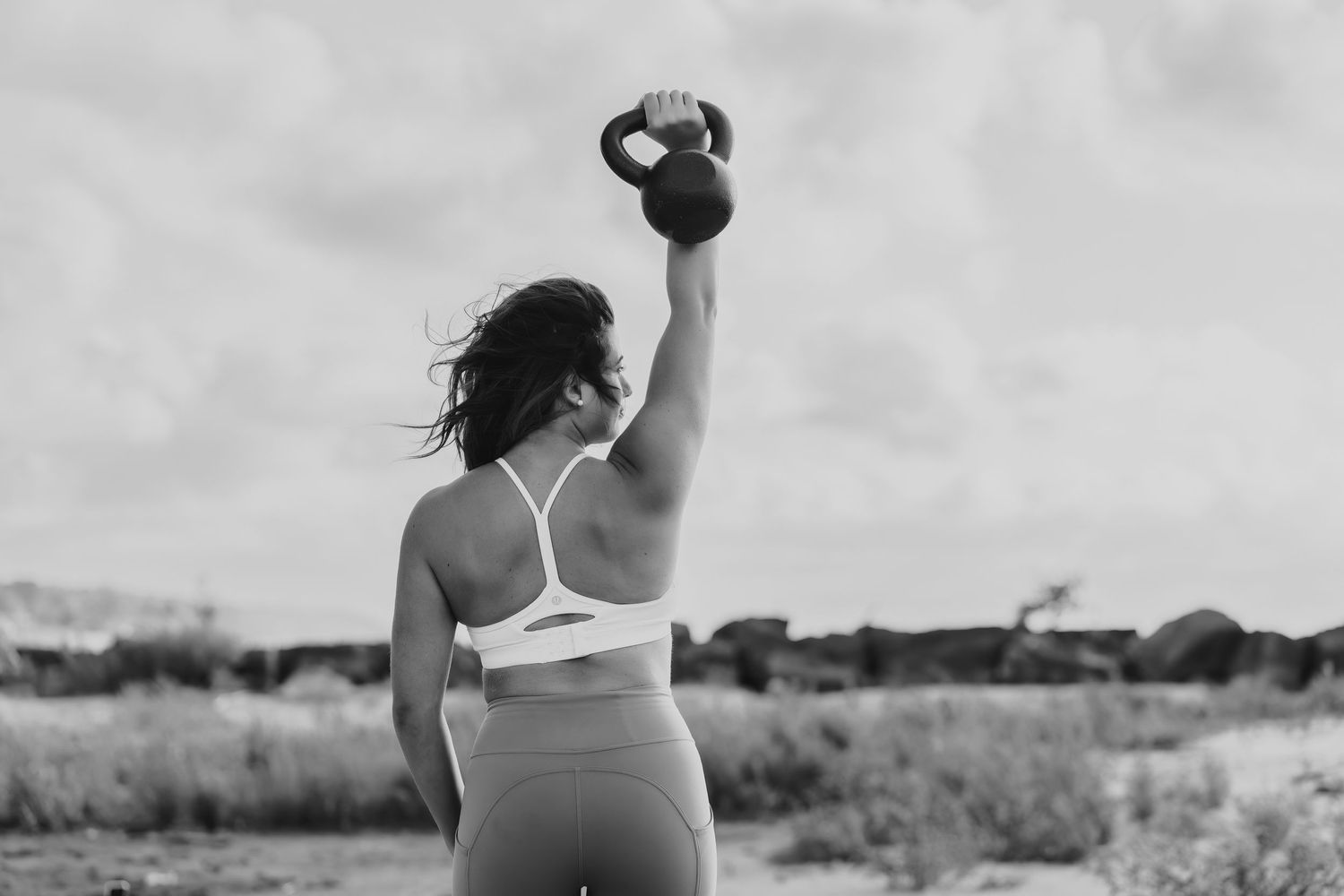 Athletic figure performs overhead kettlebell press on beach in black and white photo.