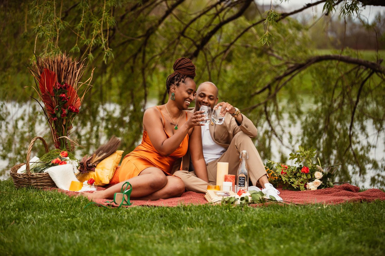 Romantic lakeside picnic scene with drinks and red floral arrangements.