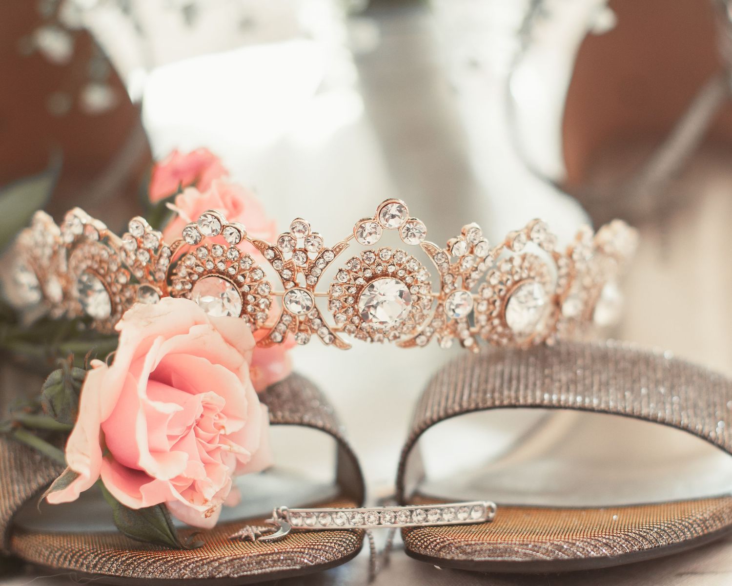 Rose gold wedding crown with crystals displayed next to a pink rose and metallic shoes.