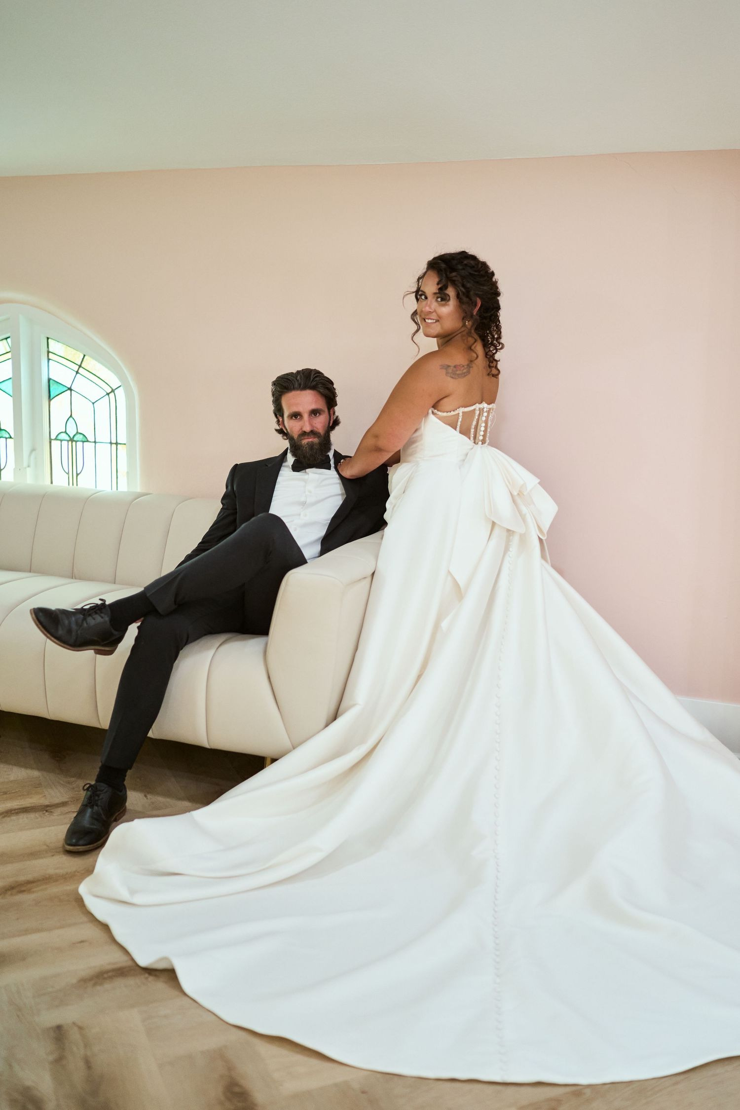 A romantic bridal couple poses together on a cream colored sofa beneath an arched window.