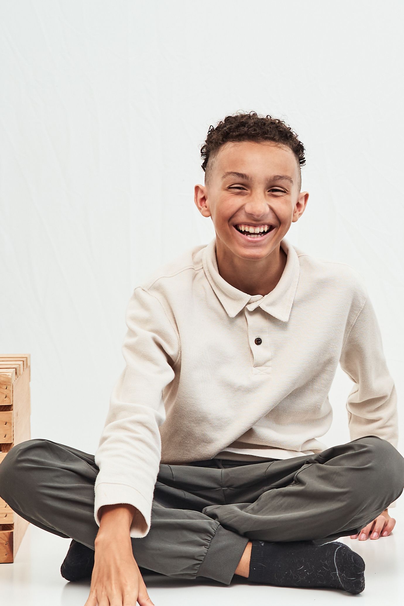 A person in a cream colored polo shirt sits cross-legged on the floor with a bright, genuine smile.