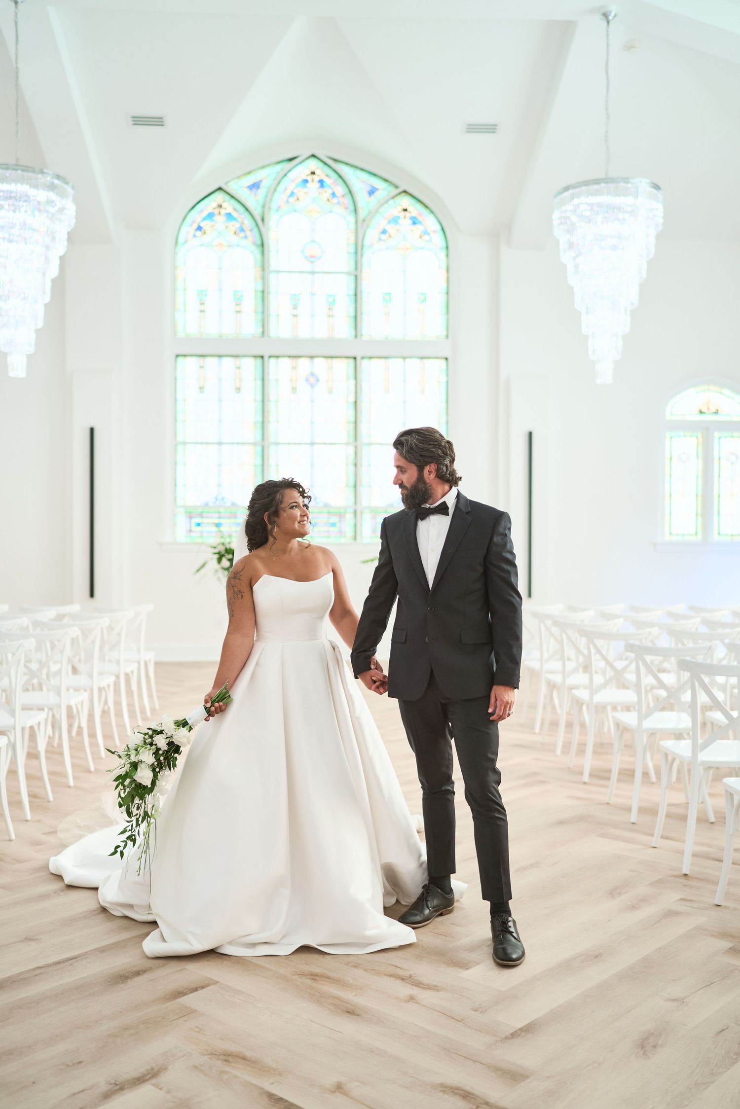 Couple in wedding attire shares intimate moments in a bright white chapel with arched windows and rustic wooden floors.