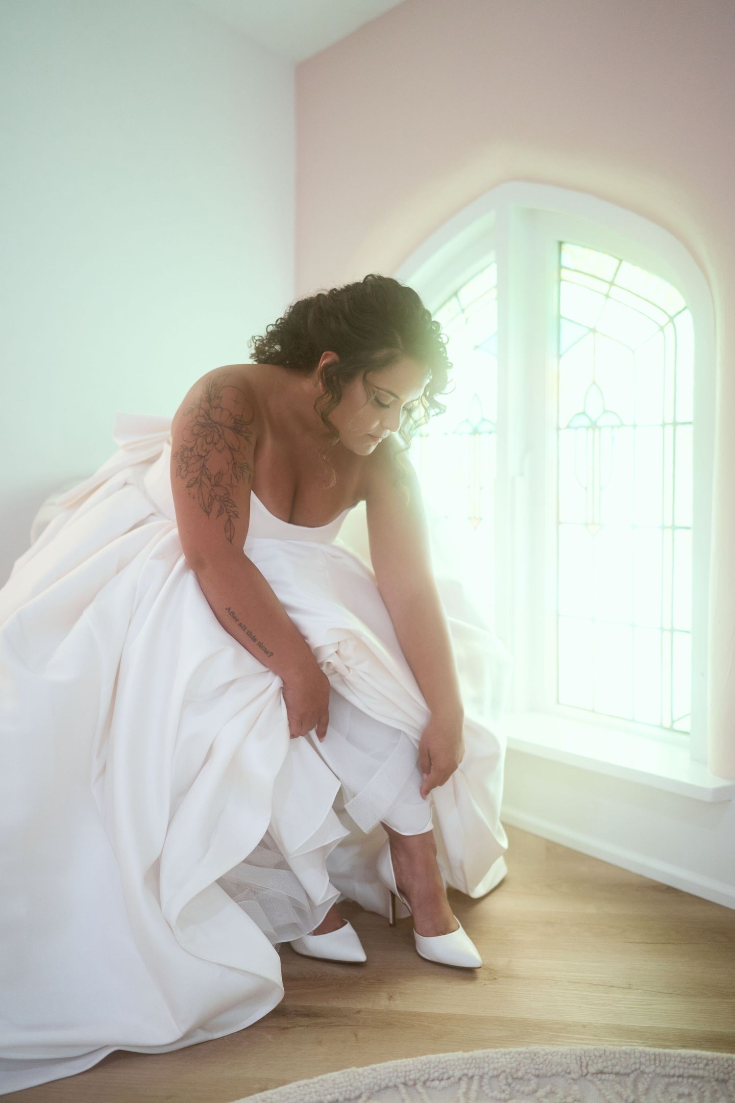A bride in a white gown adjusts her shoe near a sunlit arched window.