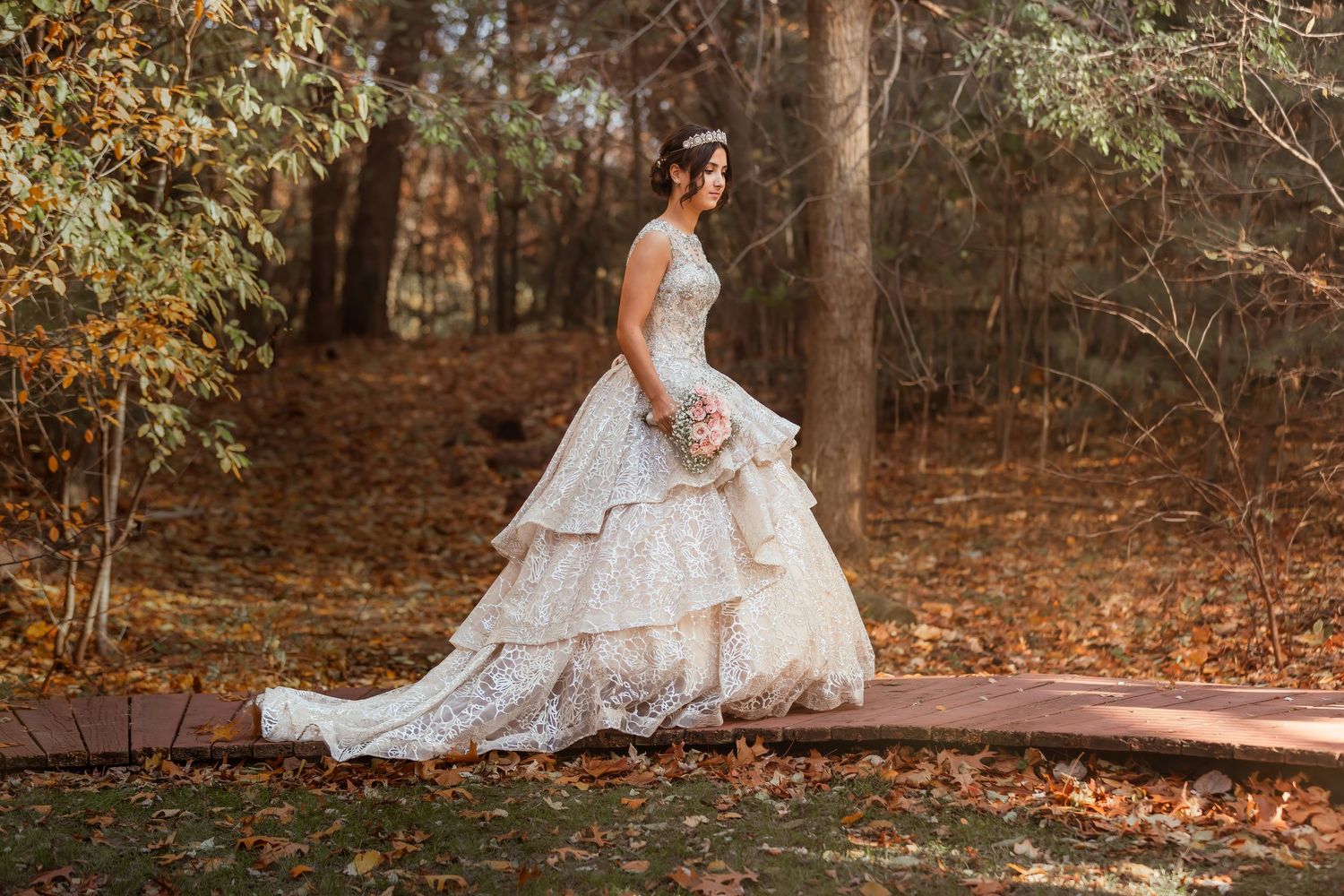 A bride in a flowing white lace gown poses gracefully among autumn leaves in a woodland setting.