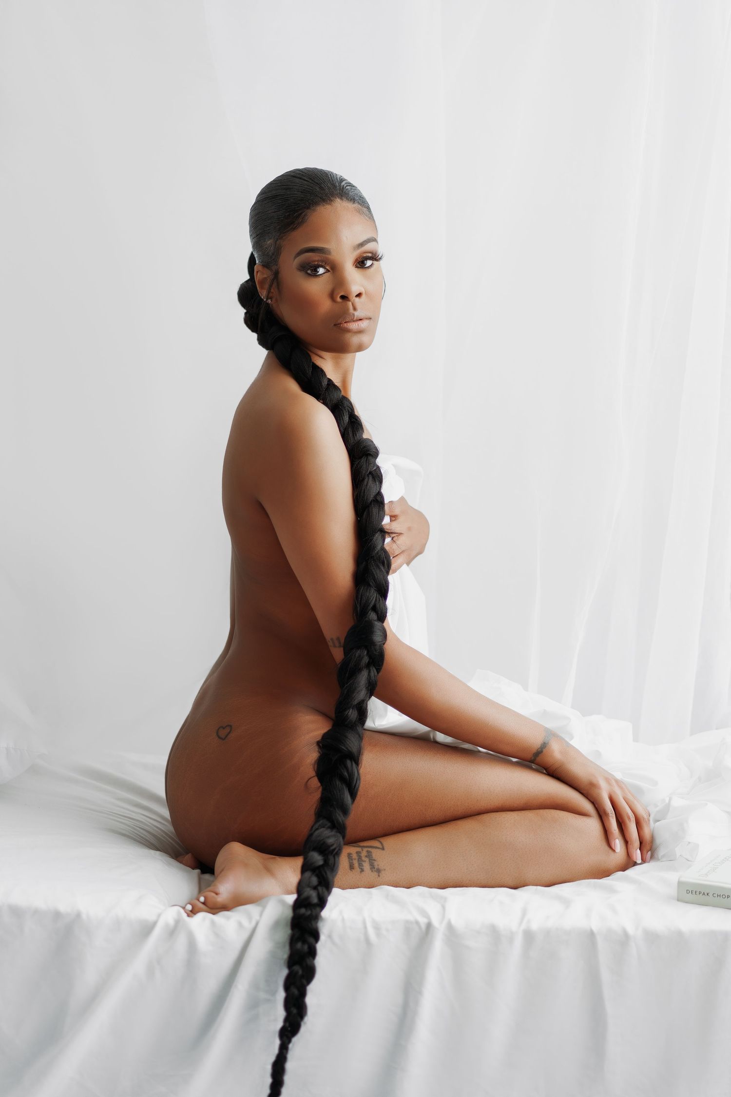 Artistic nude portrait of a woman with long braided hair sitting on white fabric in minimalist studio setting.