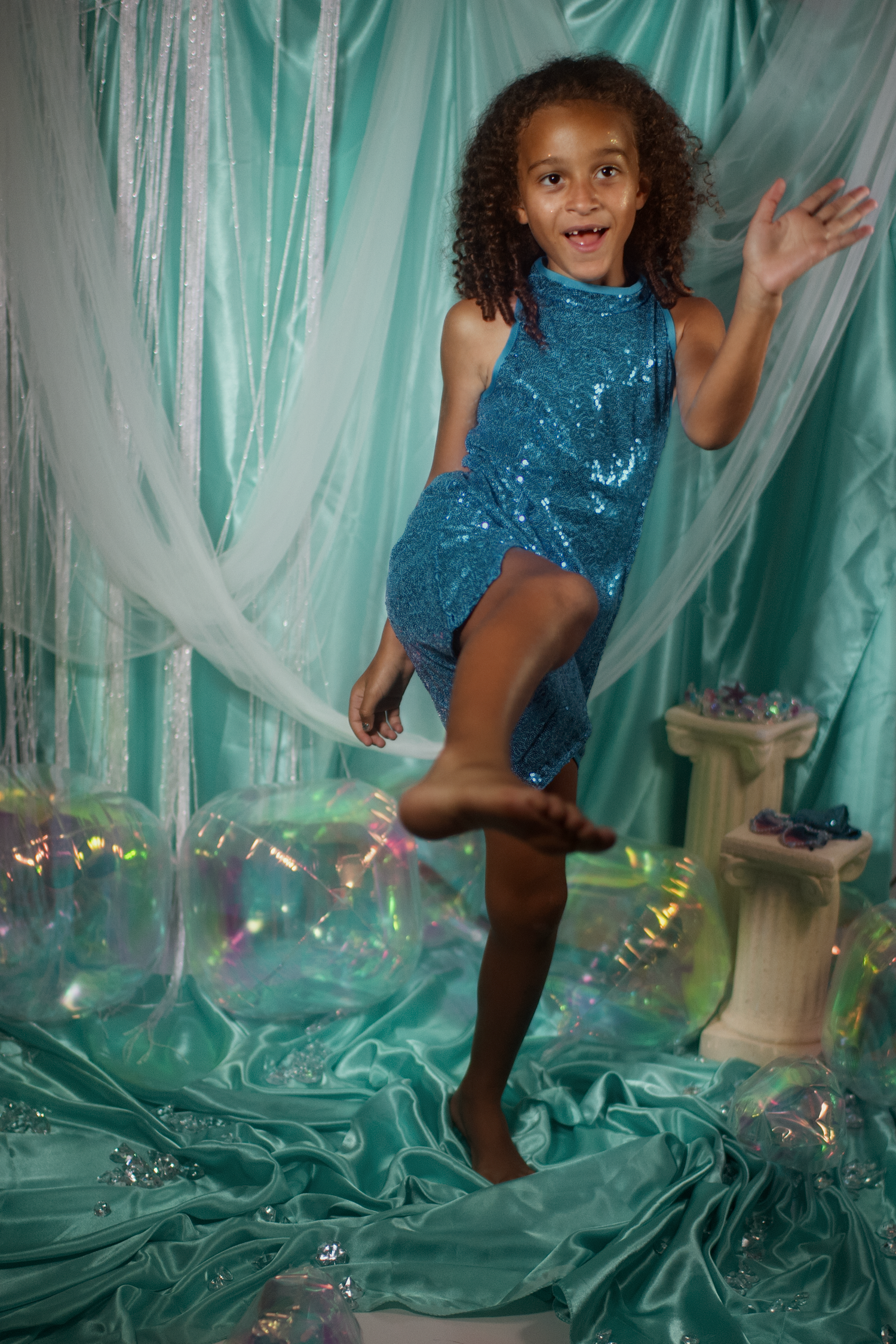 A young girl in a teal sequin dress poses barefoot, lifting one leg, surrounded by sheer aqua fabric and bubbles.