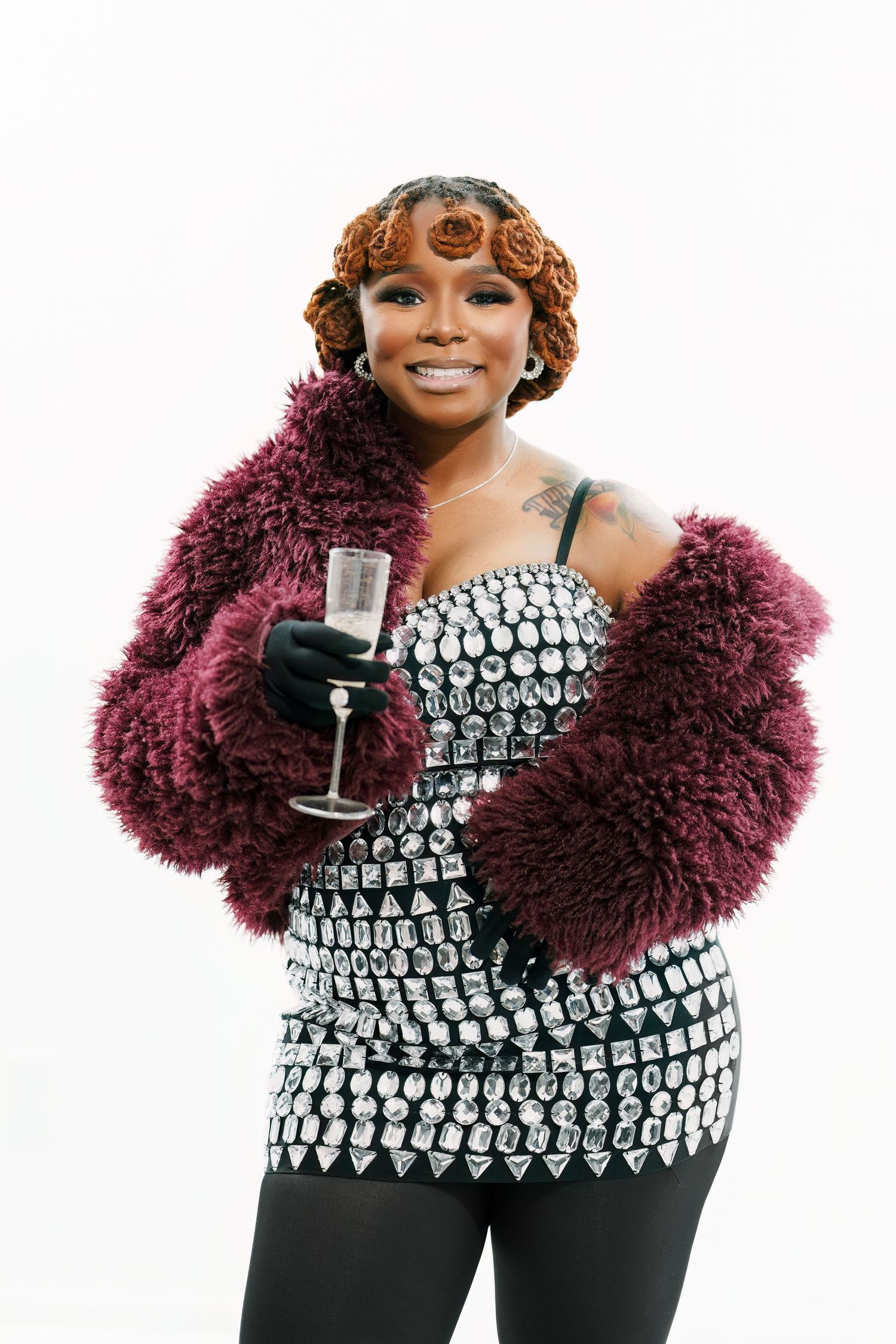 Glamorous model in sequined top and burgundy fur coat holds champagne glass against white backdrop.