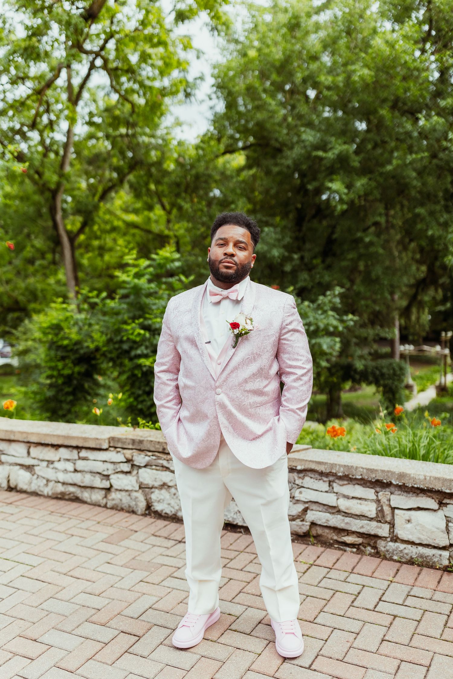 Groom in light pink suit and bow tie stands on stone patio surrounded by lush greenery.