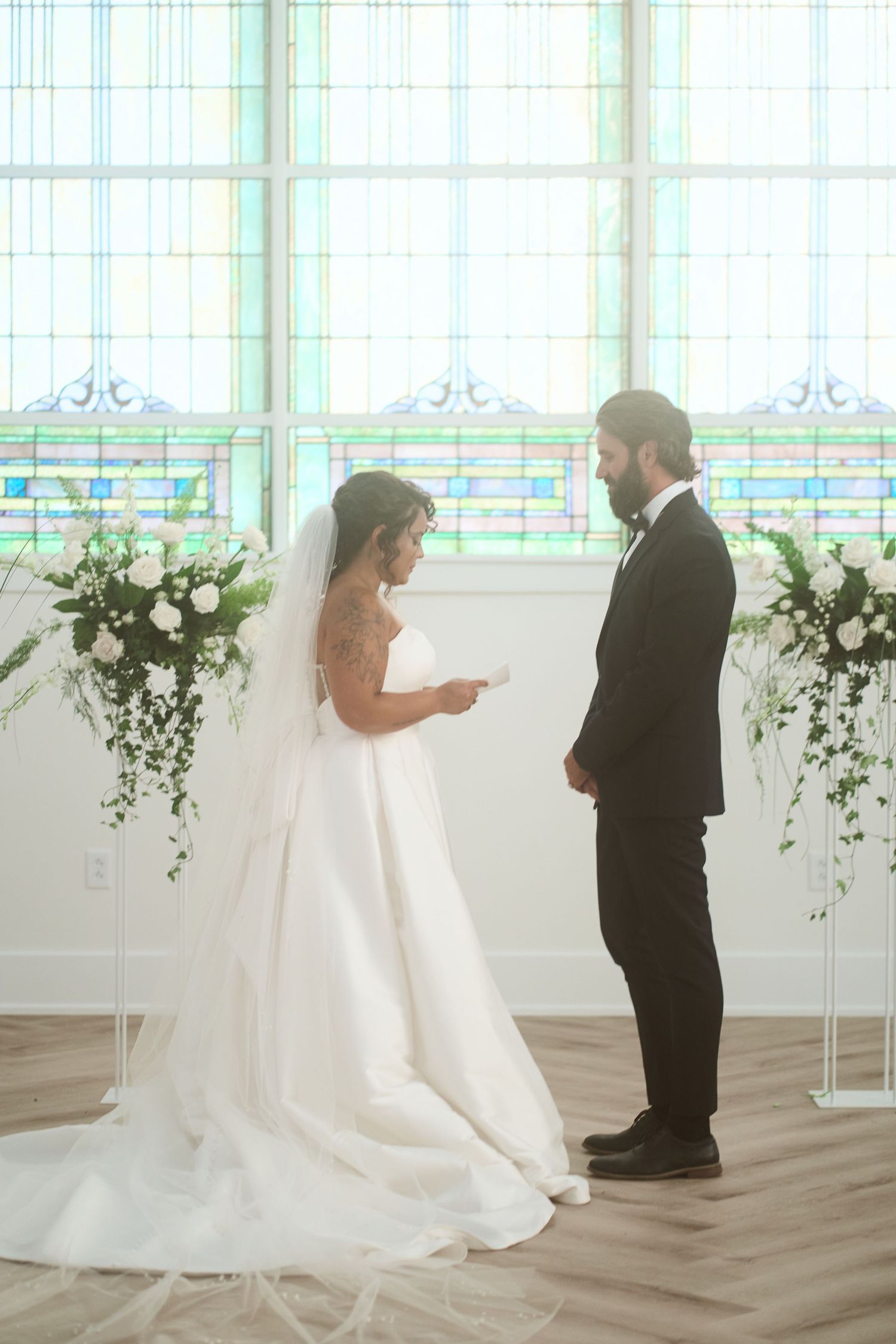 A couple shares a private first look moment in an elegant white chapel with arched windows and floral decor.