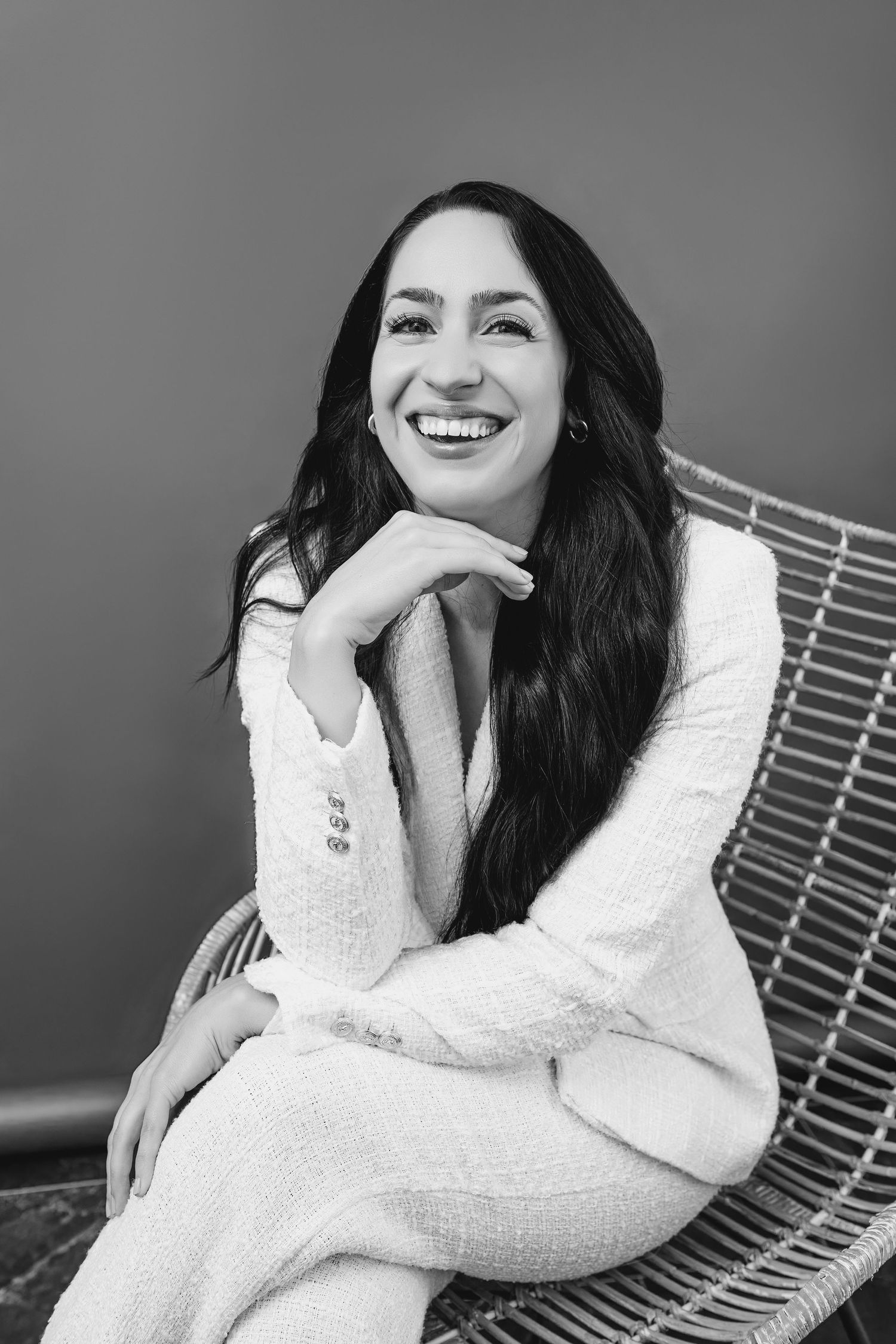 Black and white portrait photo of someone in a white sweater sitting on a chair with a joyful smile.