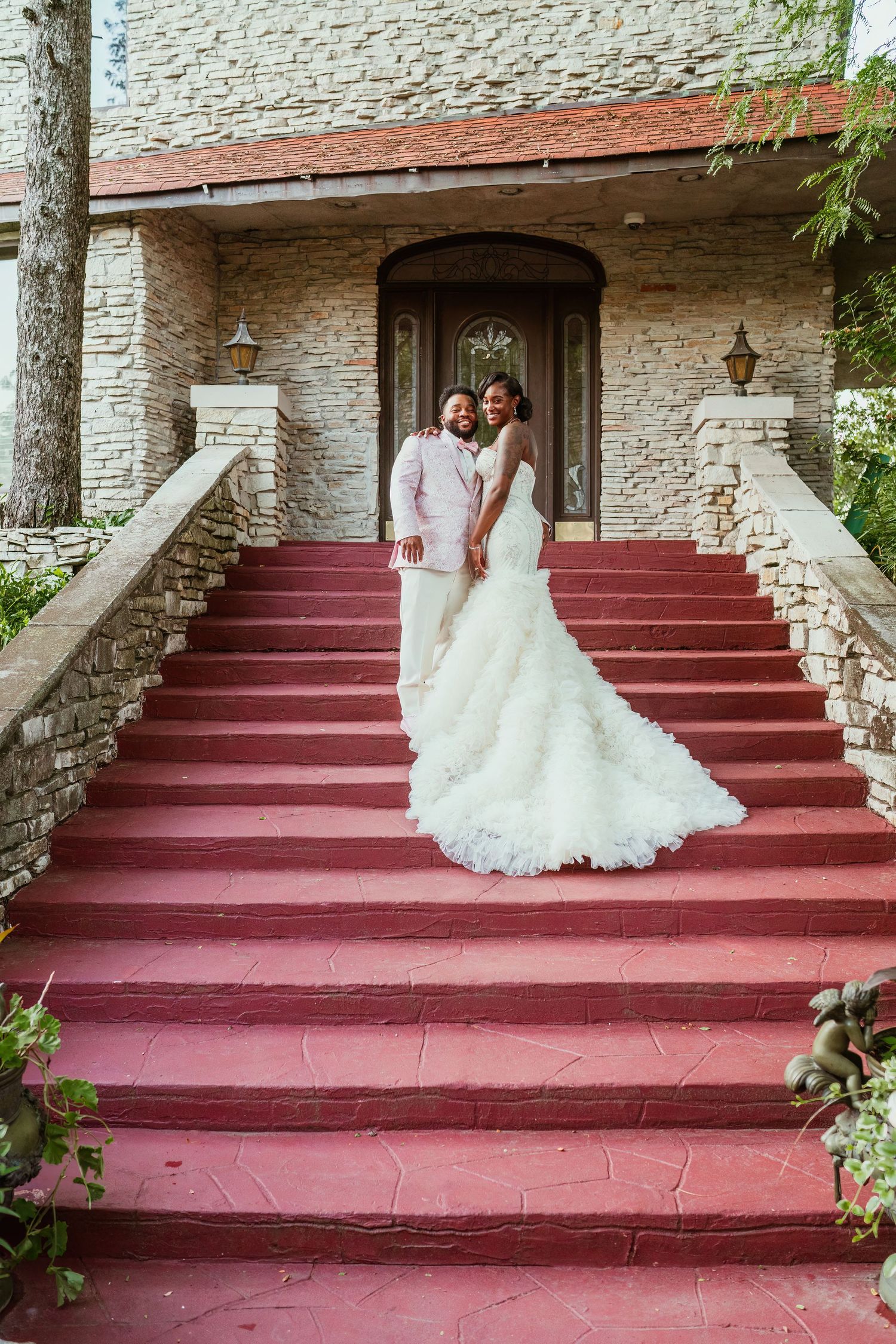 A wedding couple stands on red stairs against a stone facade with elegant landscaping.
