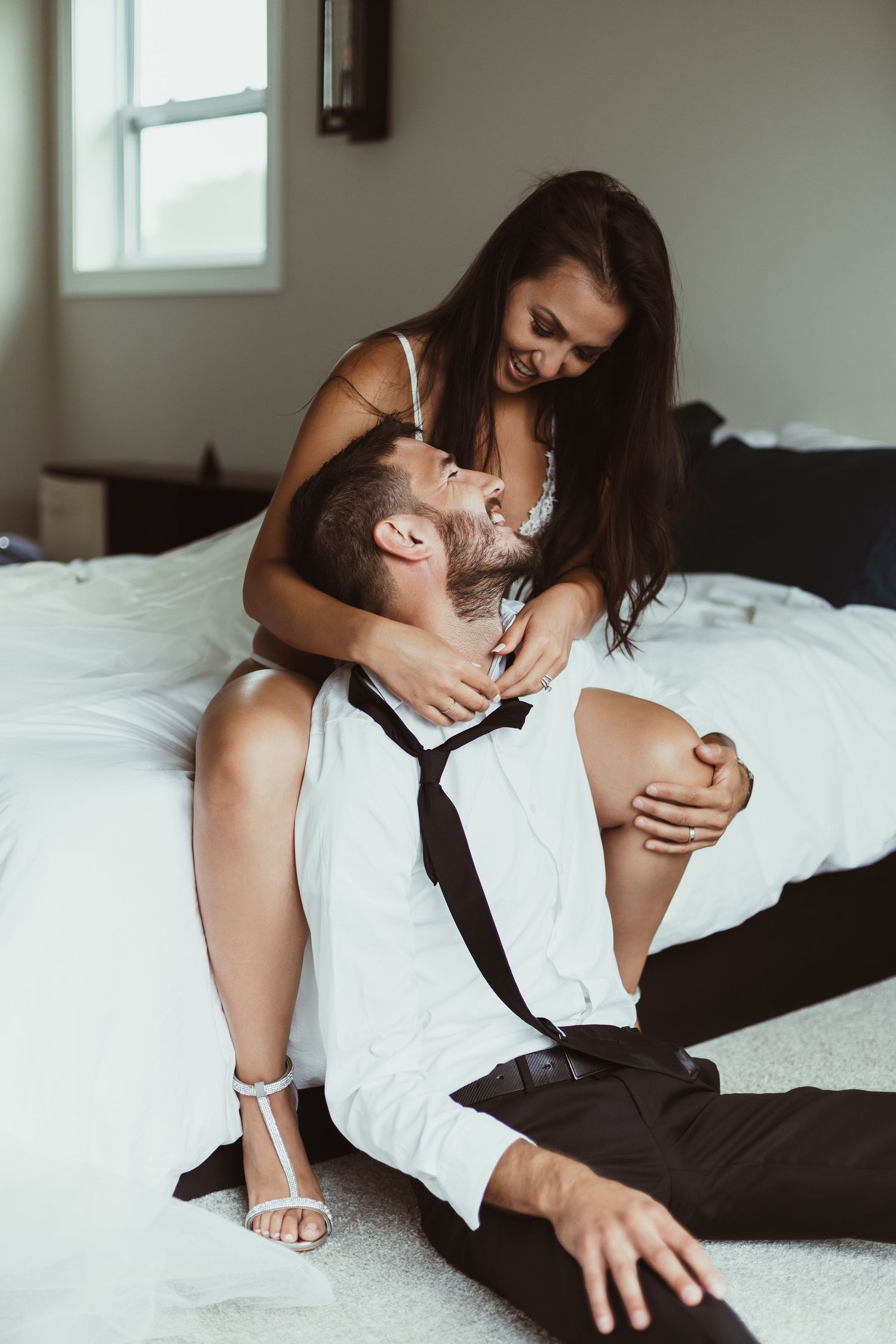 Playful boudoir photo of couple on white bedding in formal wear.