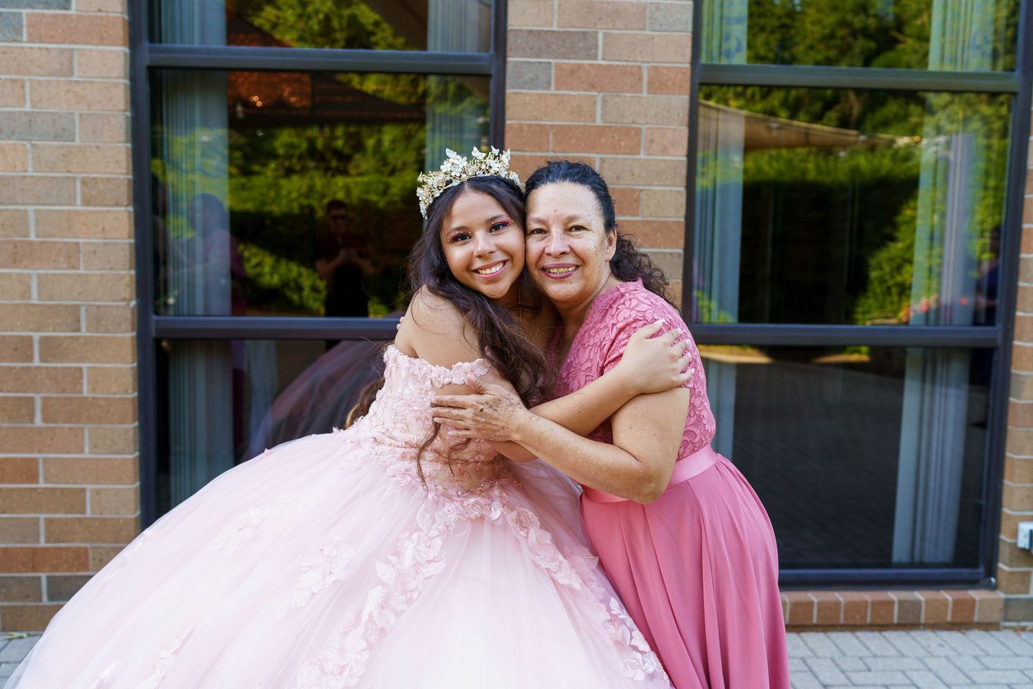 Two people embrace while wearing formal dresses in pink tones outside a brick building.