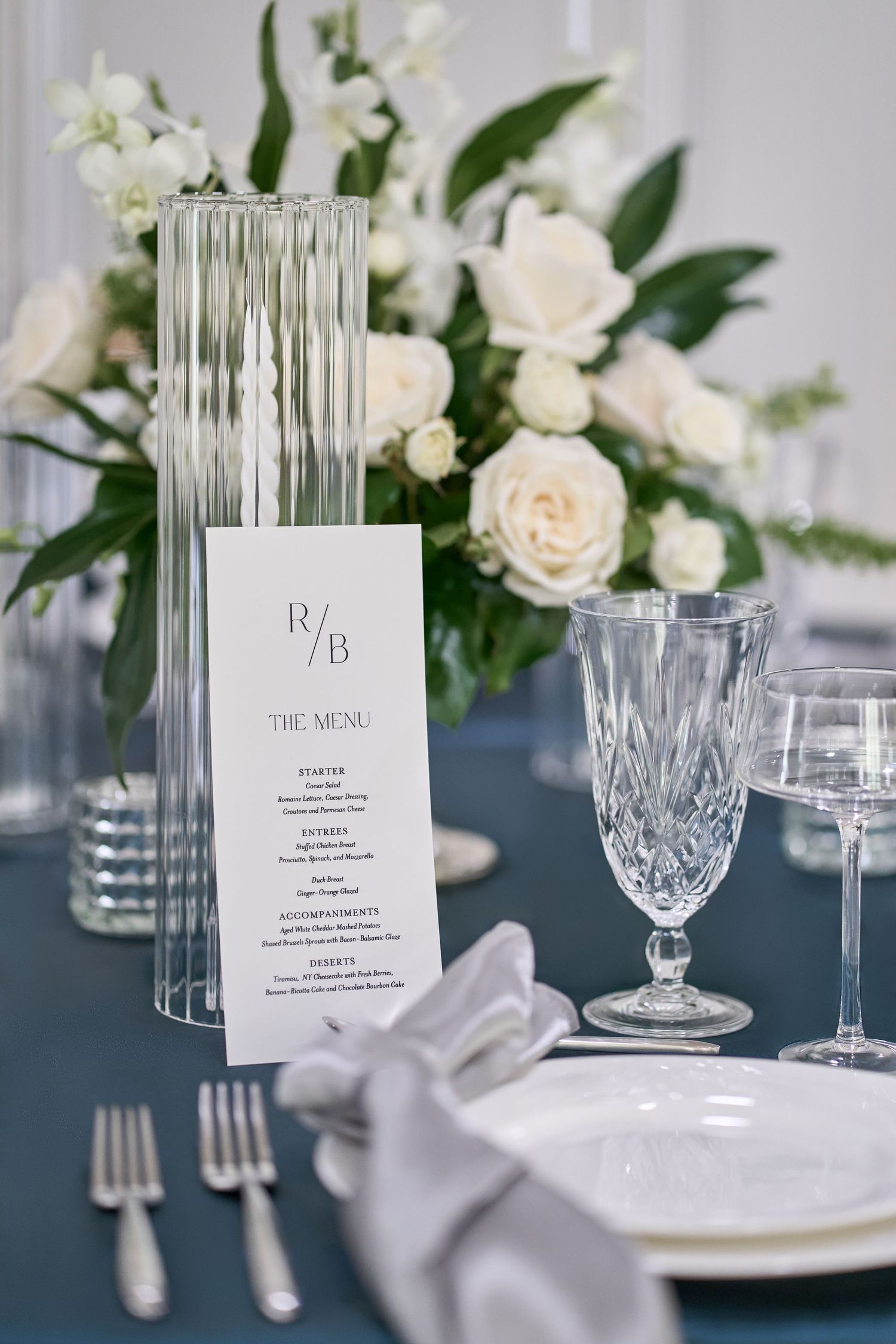 Elegant wedding table setting with crystal glassware, white roses, and formal menu card on navy blue tablecloth.