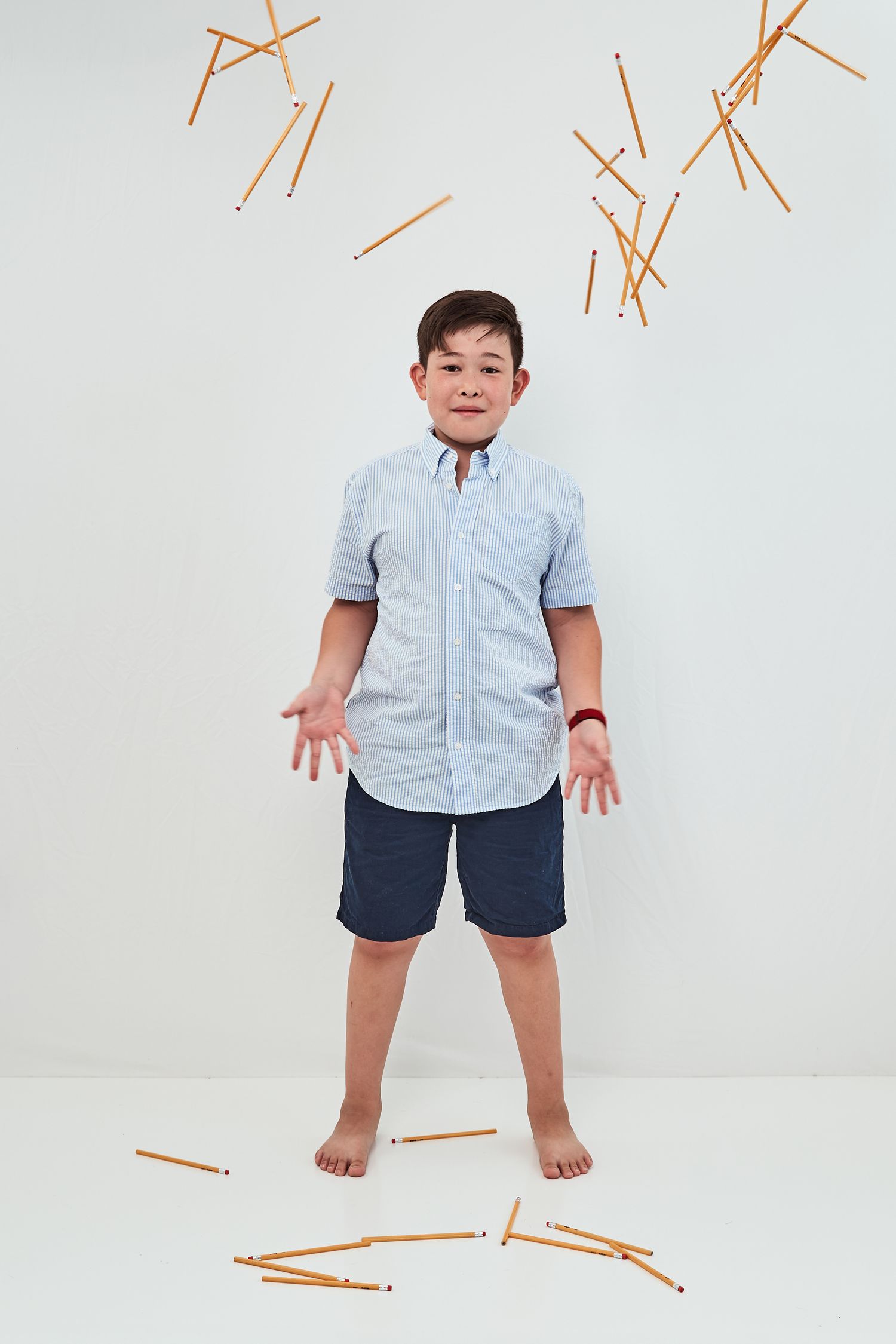 A young person stands barefoot against a white wall with orange sticks scattered around in a casual summer outfit.