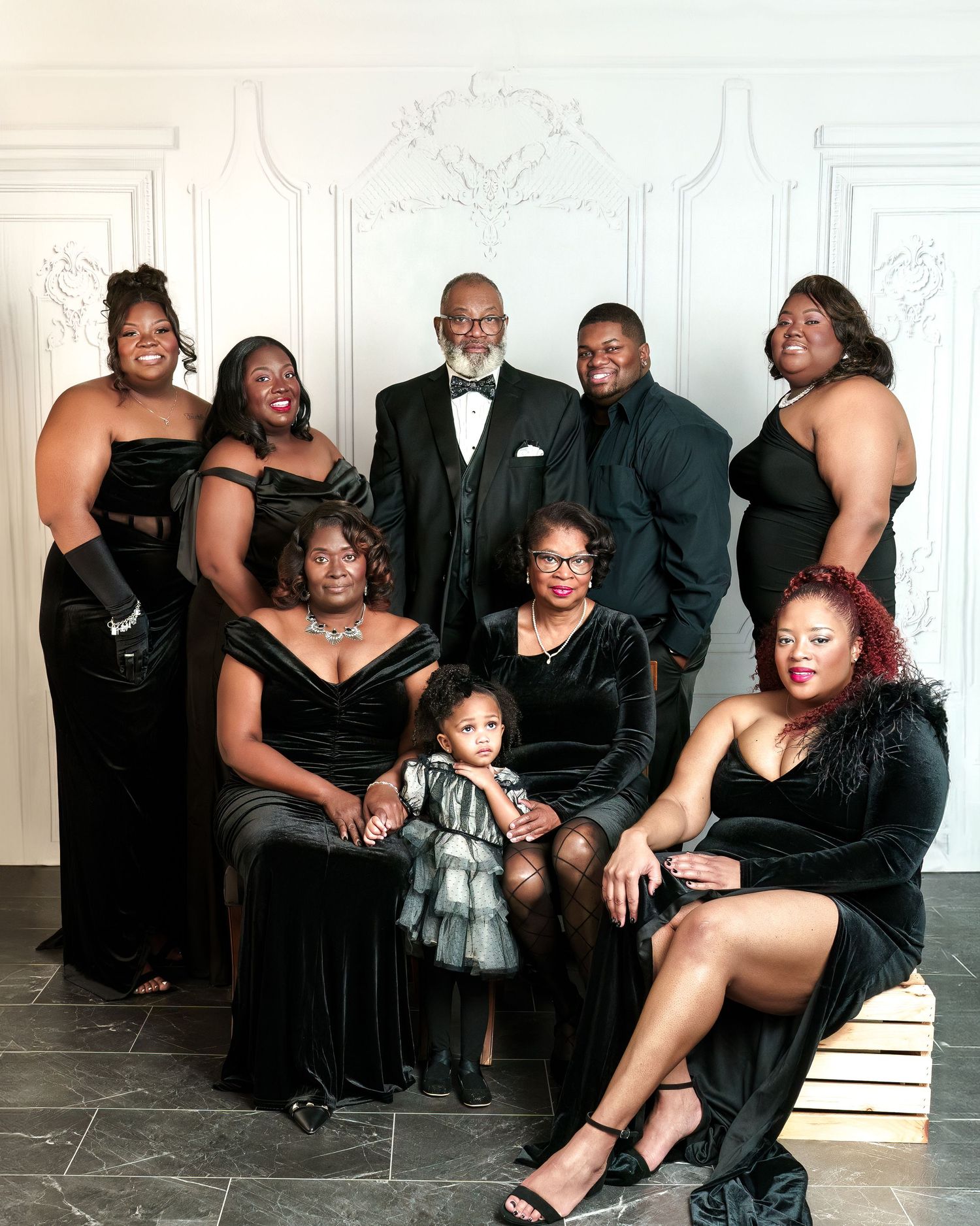 A formal family portrait in black and white attire against an ornate white wall with elegant decorative molding.
