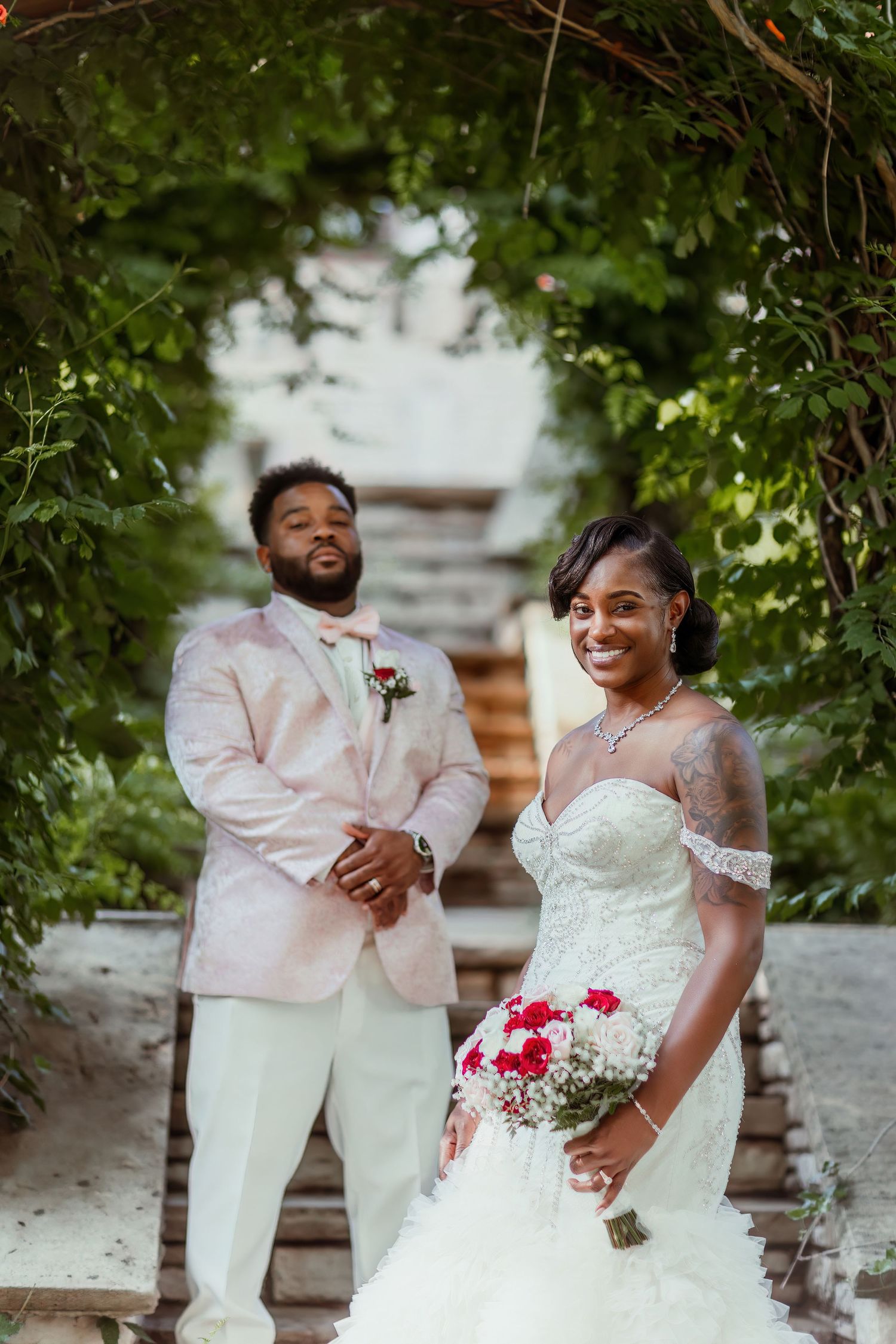 A newlywed couple poses beneath a leafy garden archway on stone steps.
