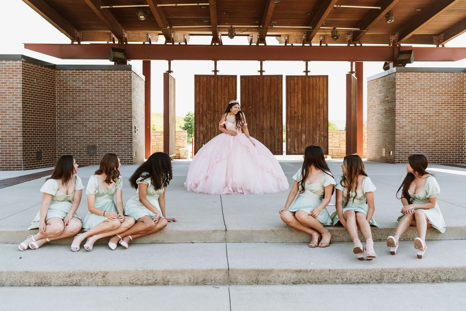 A group in pastel dresses gathers under a wooden pergola during a celebratory outdoor photo session.