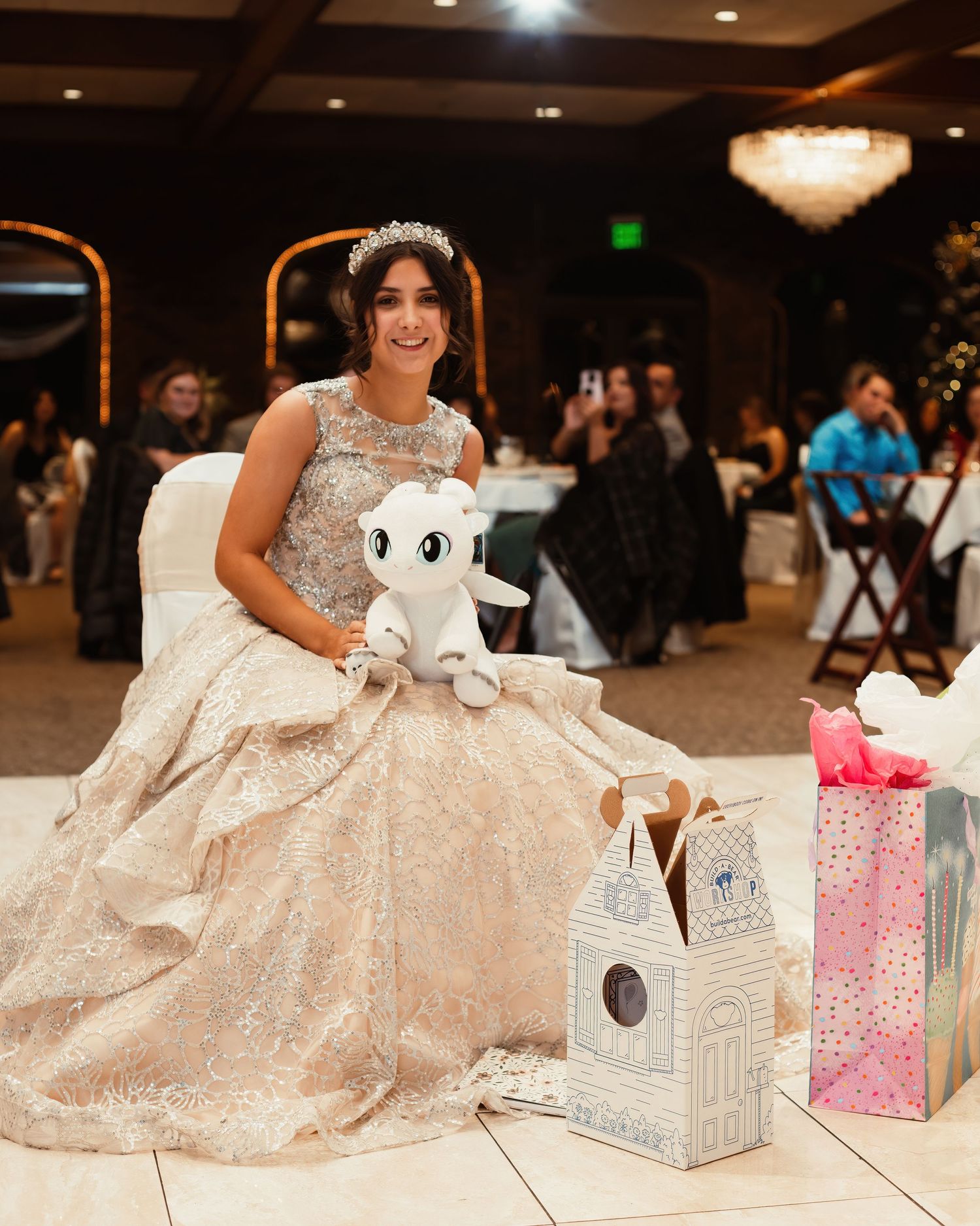 A bride in a sparkly ballgown poses with a white plush toy and decorative gift boxes at her reception.