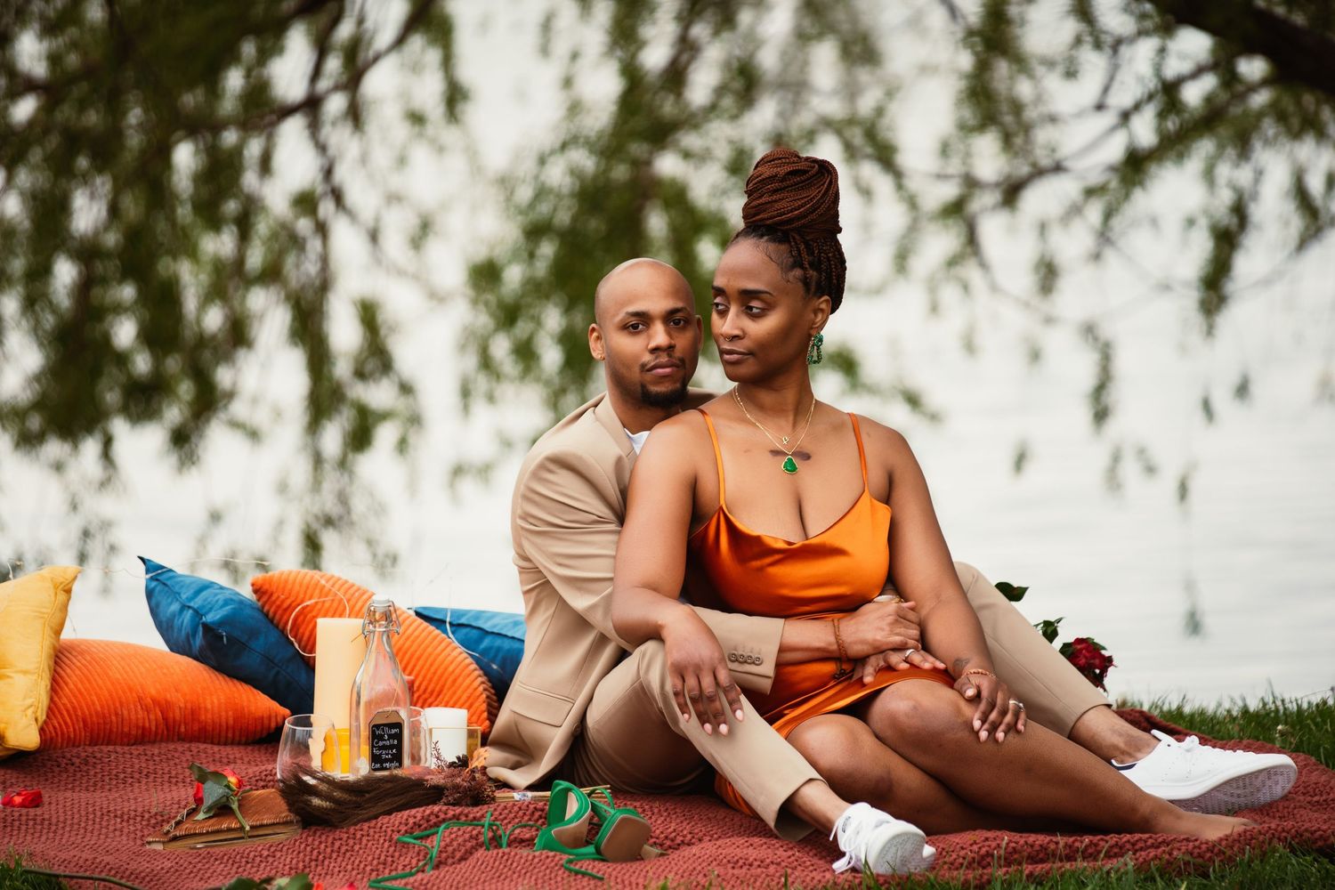 Couple enjoying a romantic outdoor picnic with orange drinks and blanket by a lake.