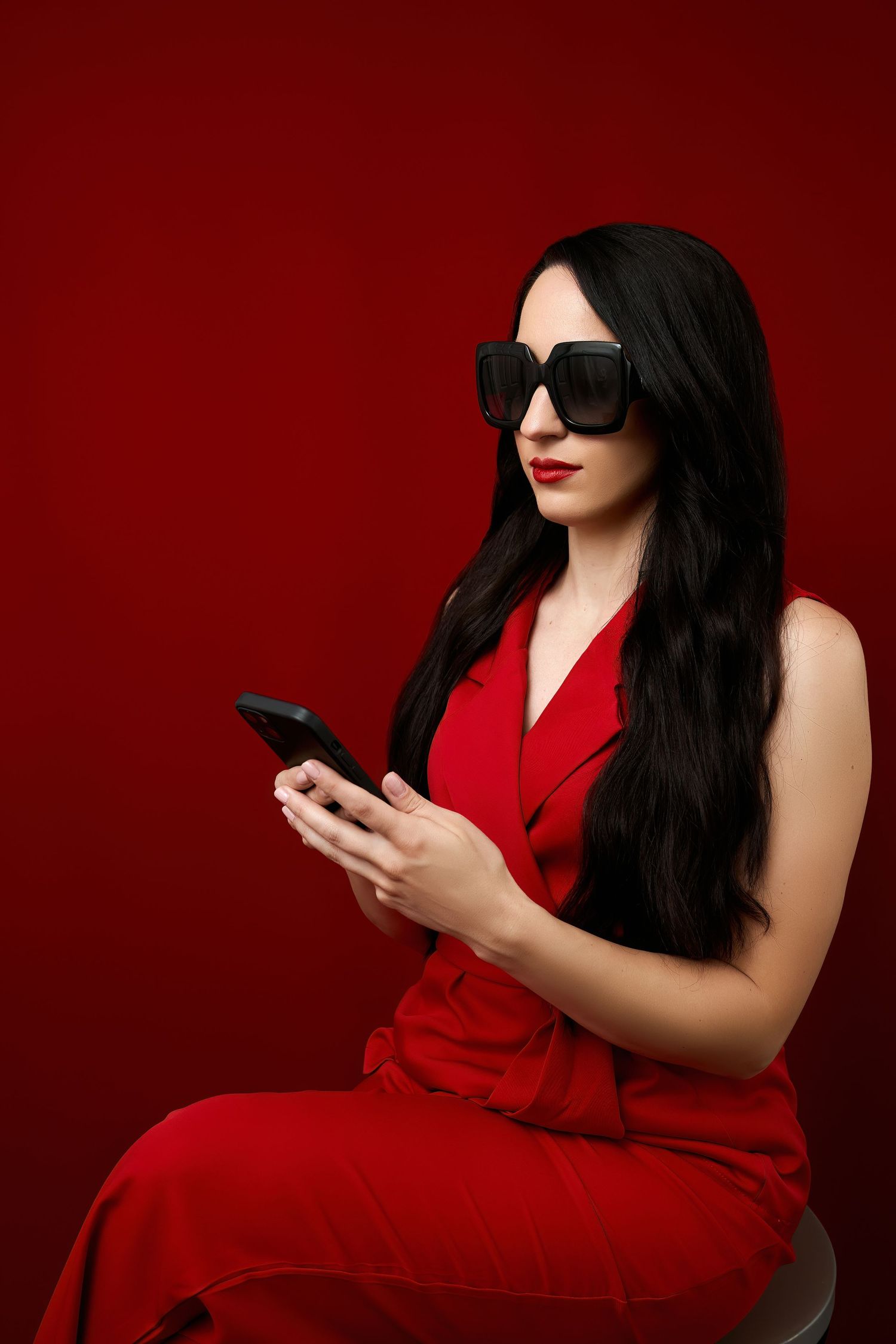 A stylish figure in a red dress and sunglasses sits looking at their mobile phone against a red background.