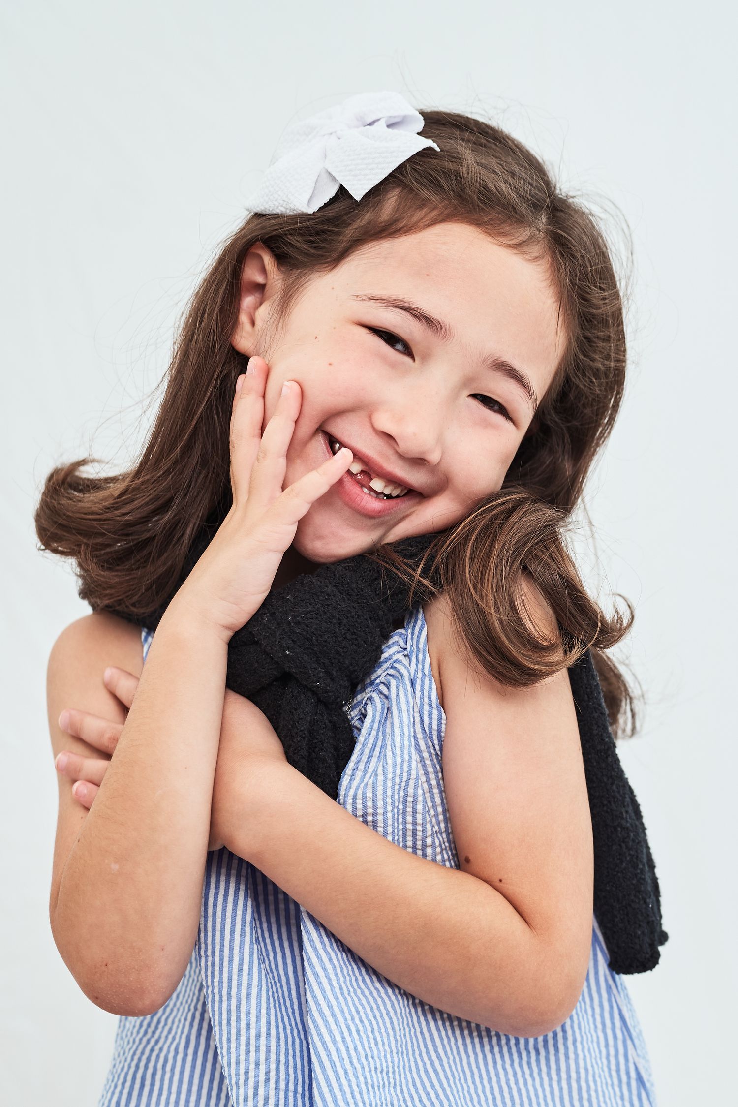 A cheerful young student in a blue striped top and white hair bow giggles while covering their smile.