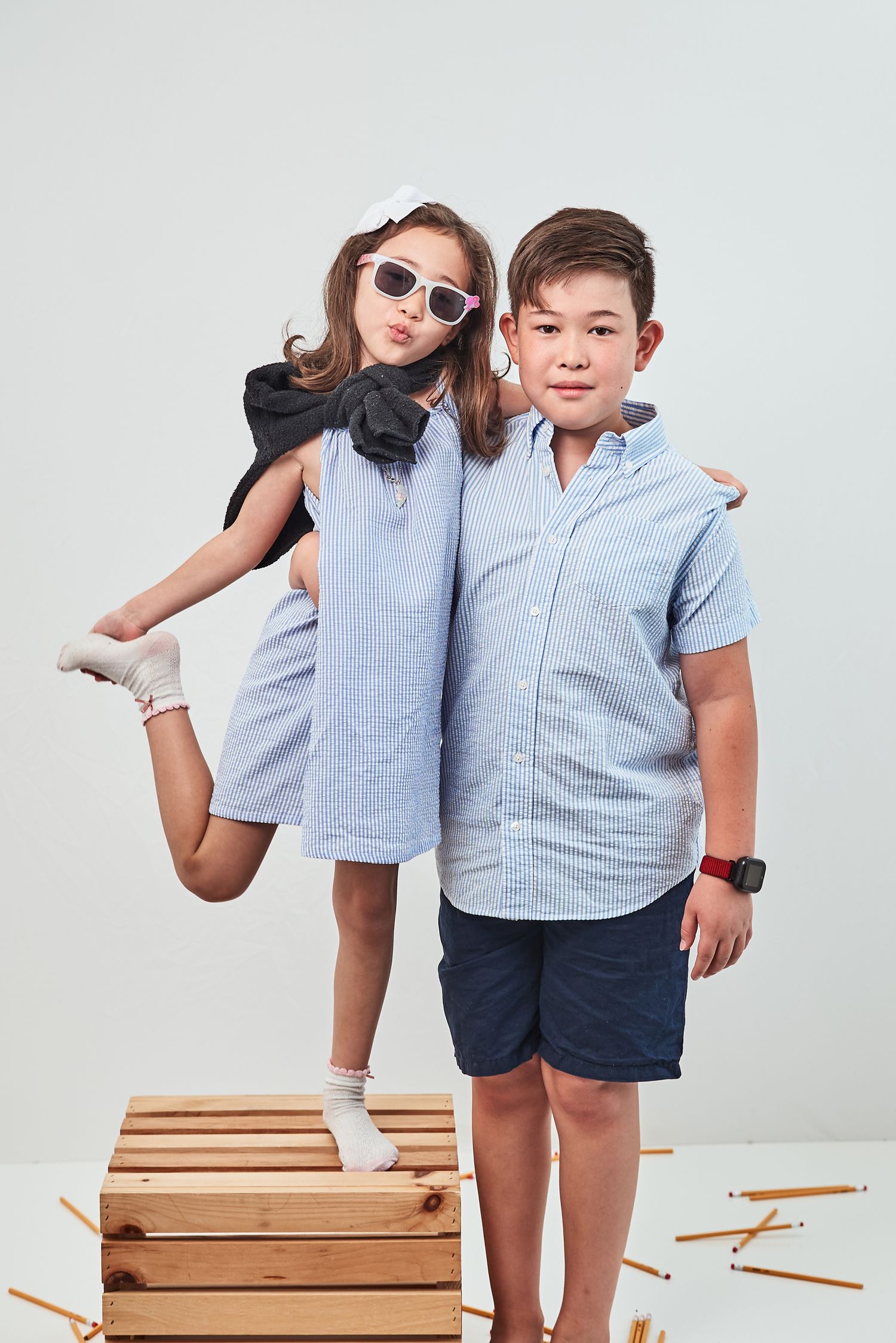 Two kids in coordinating light blue outfits pose playfully on a wooden crate against a white background.