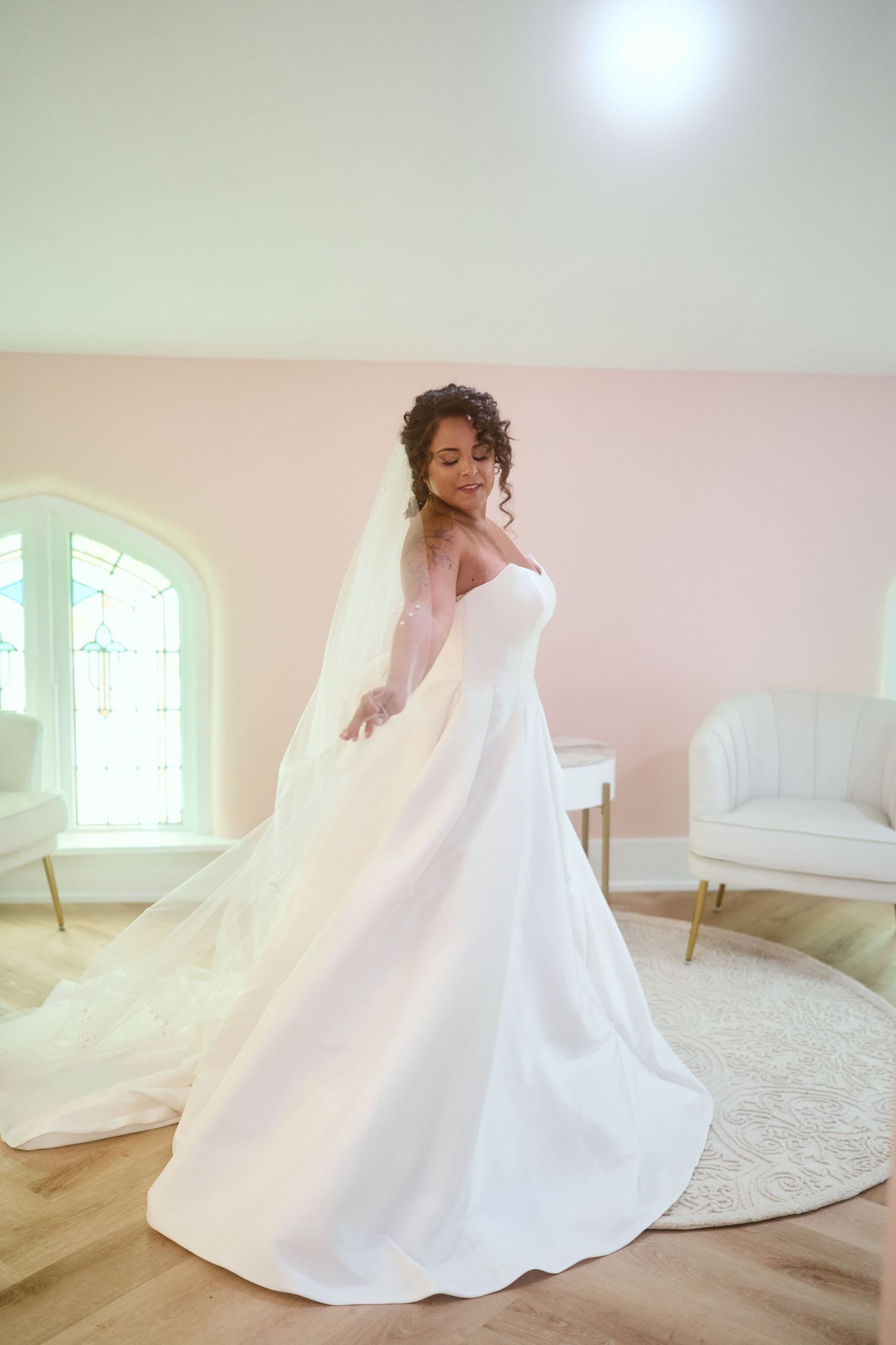 A bride twirls in an elegant white ballgown wedding dress with a flowing veil in a soft pink room.