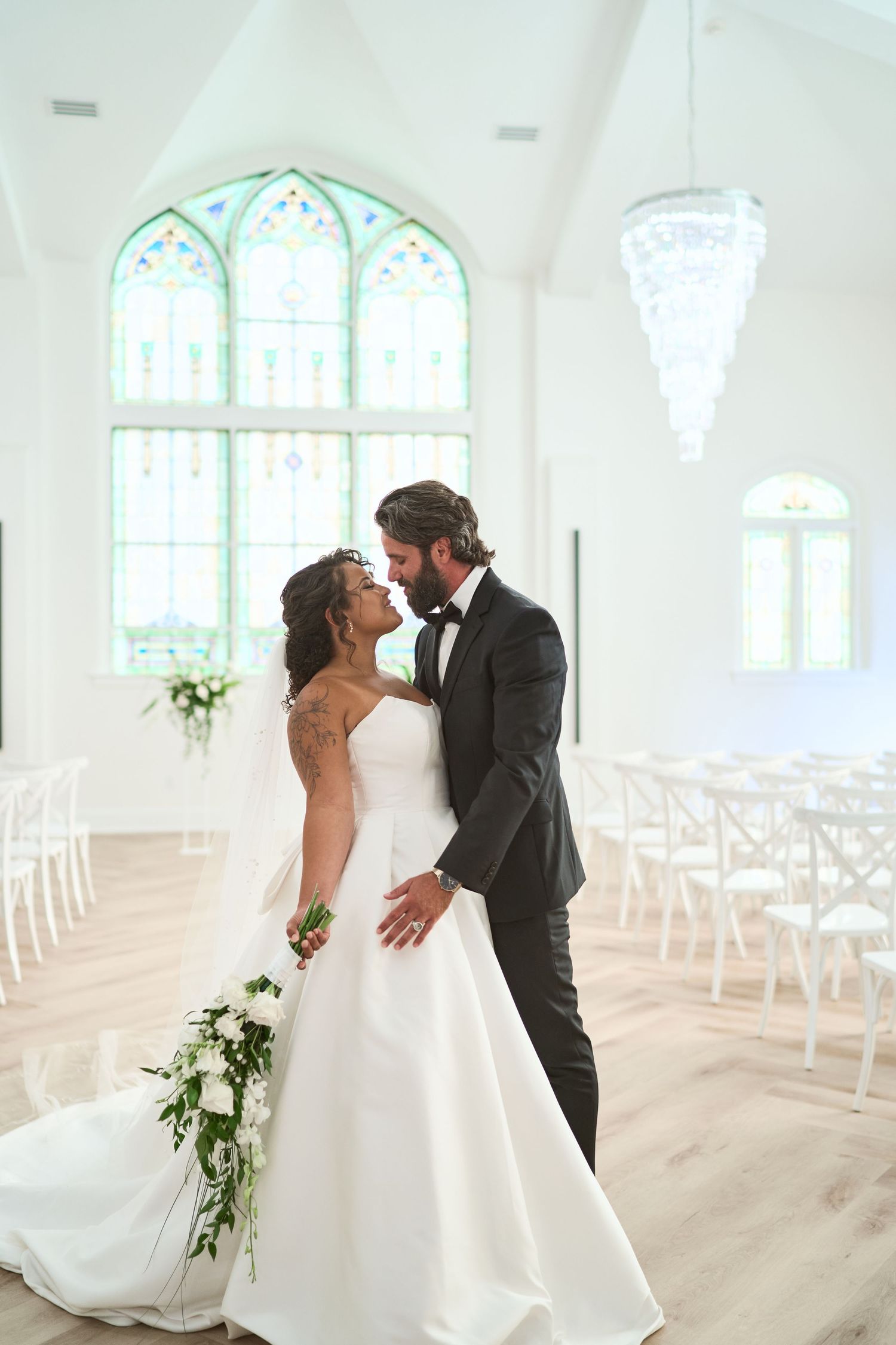 An intimate wedding moment in an elegant white chapel filled with natural light from arched windows.