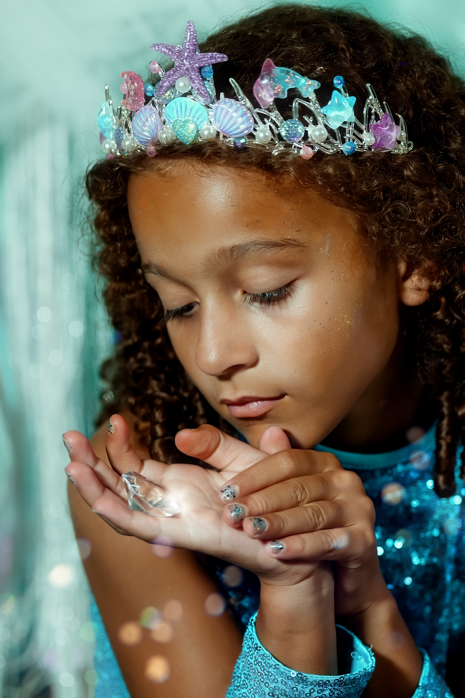 Young girl in mermaid crown and blue sequin dress holds glitter in her hands, looking down with wonder.