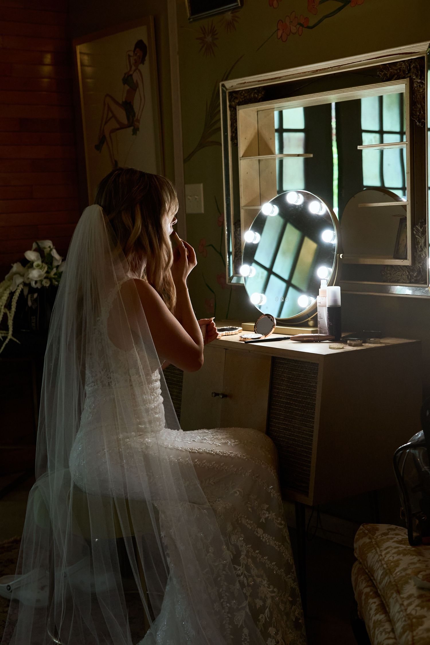 A bride in a flowing white wedding dress and veil stands near a window in moody natural lighting.