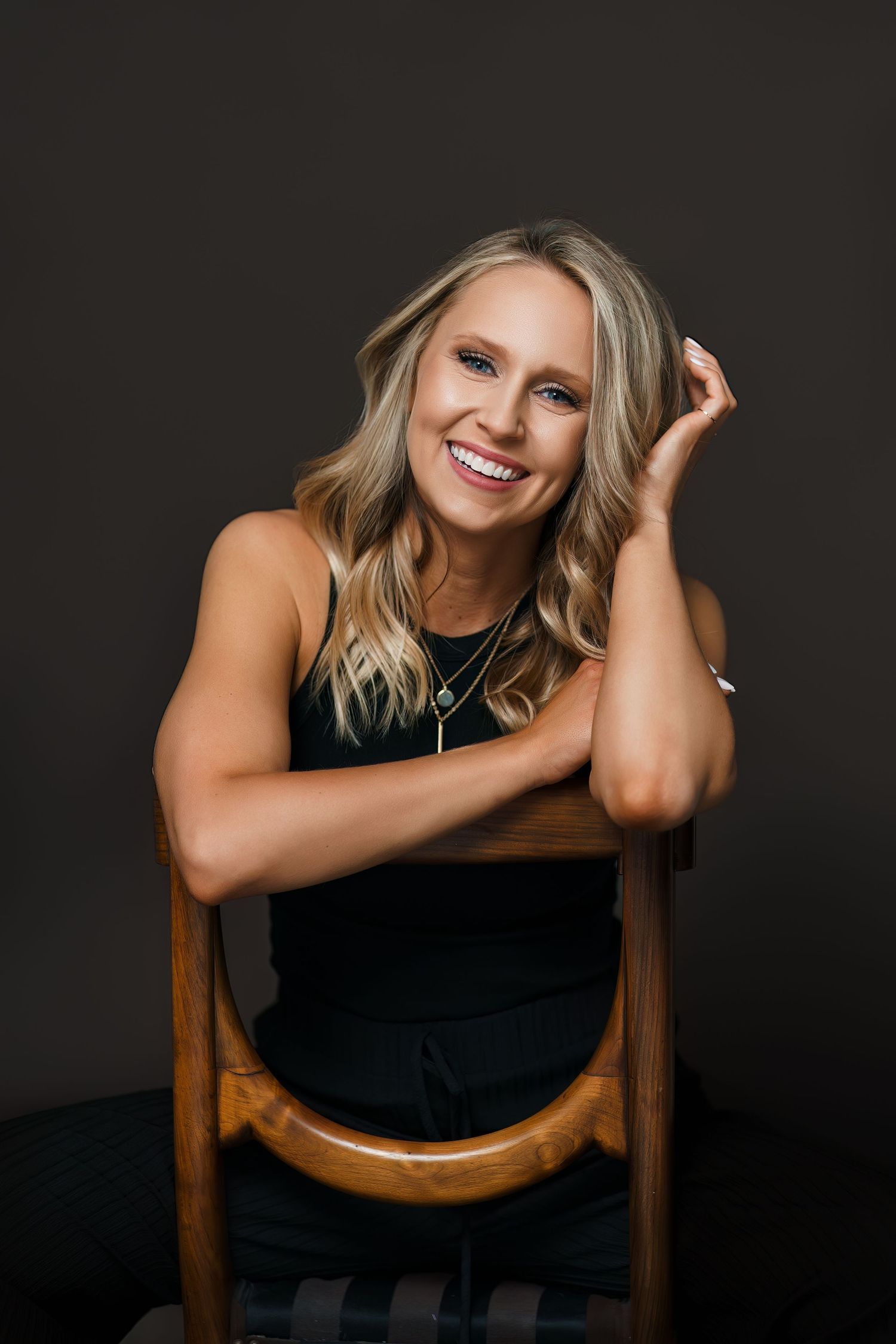 Person in black top poses playfully on wooden chair against dark background in professional studio portrait series.
