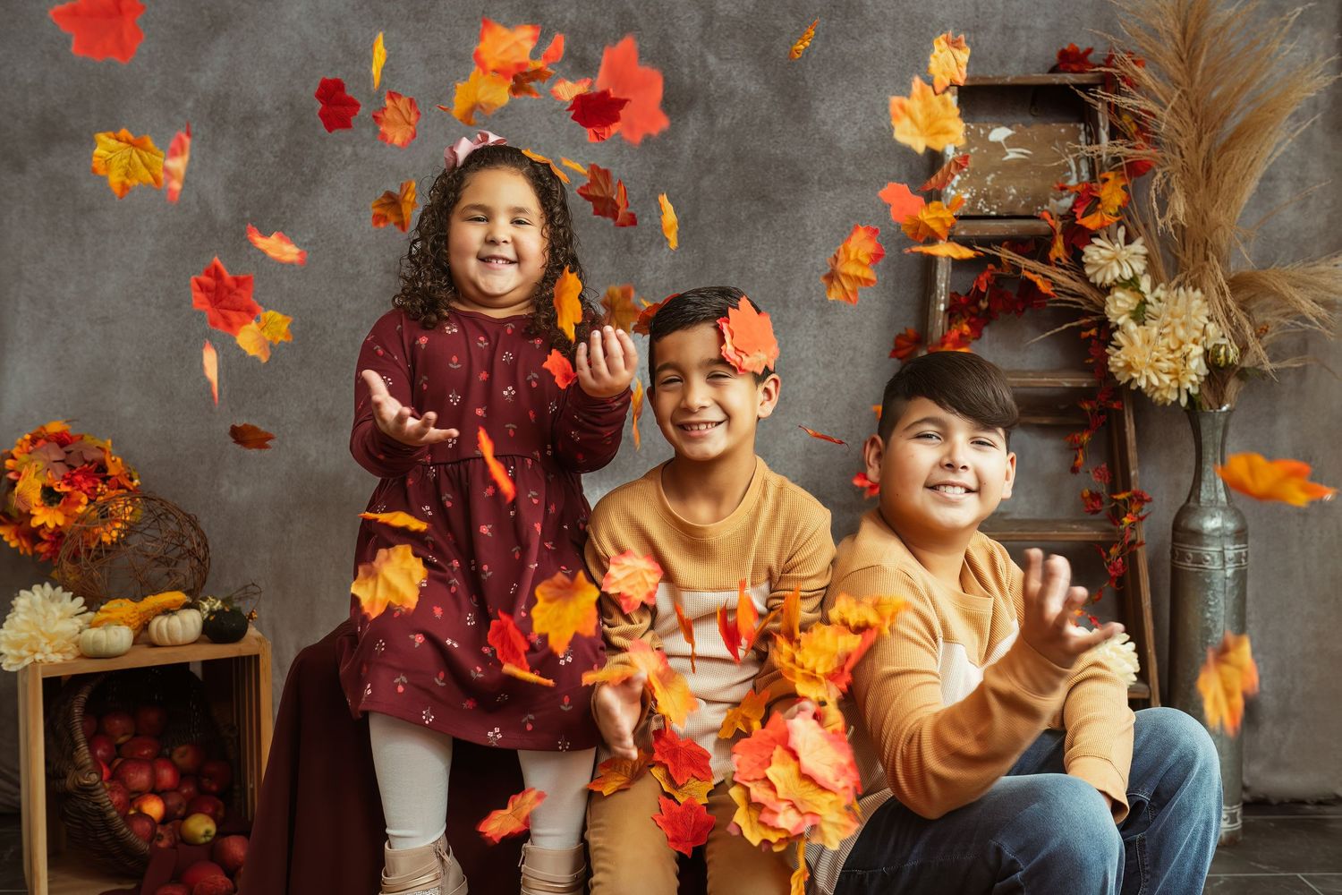 Three children play with falling autumn leaves in a cozy indoor setting with rustic fall decor and pumpkins.