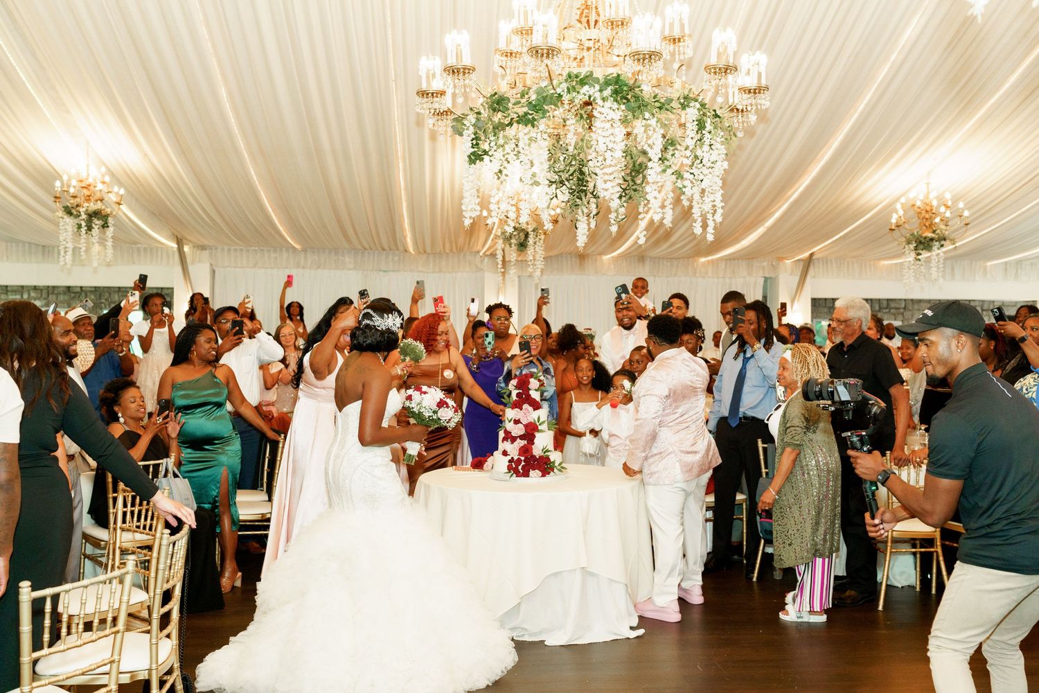 Wedding guests celebrate joyfully under an elegant chandelier decorated with cascading floral arrangements.